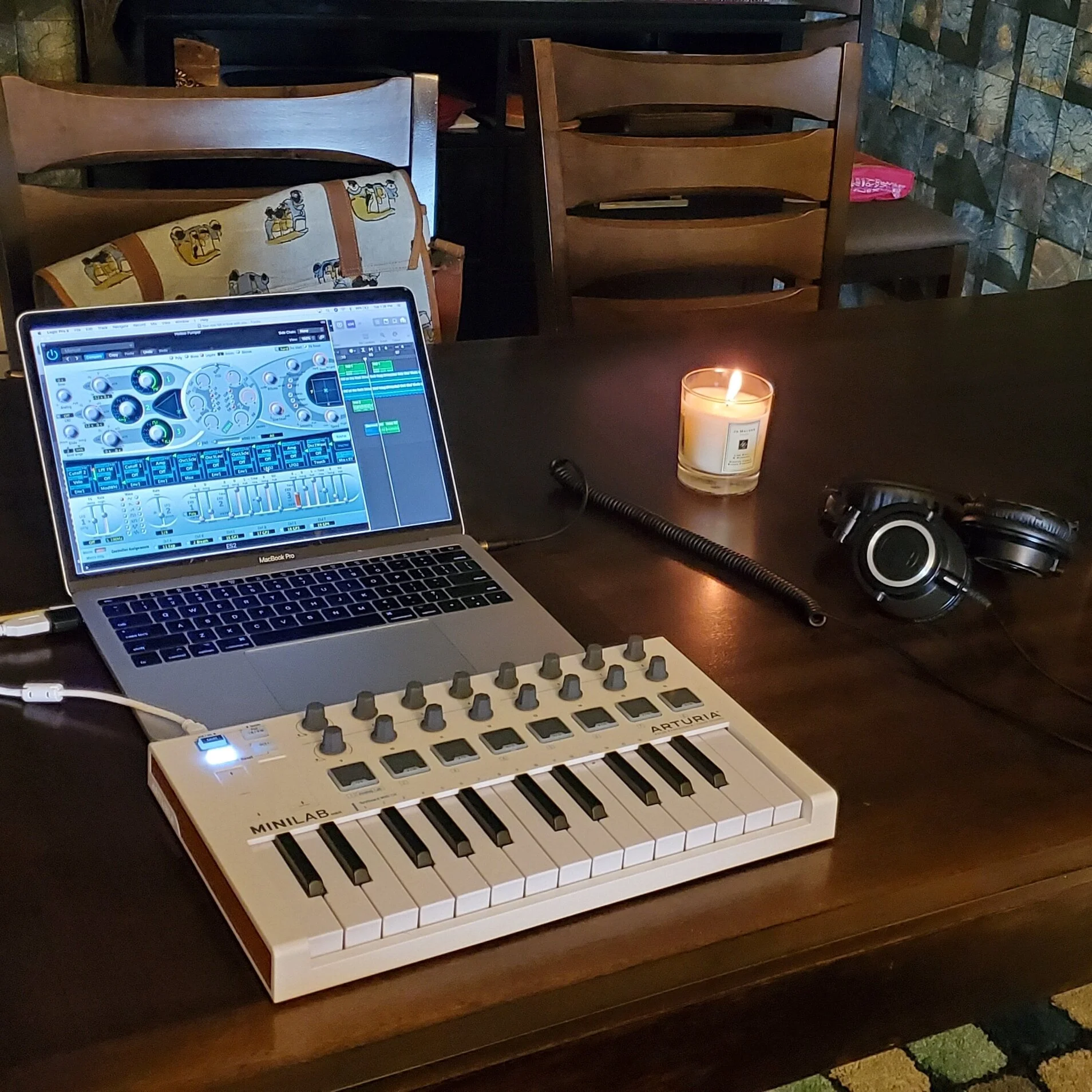 Laptop running music production software, a MIDI keyboard, headphones, and a lit candle on a wooden table.