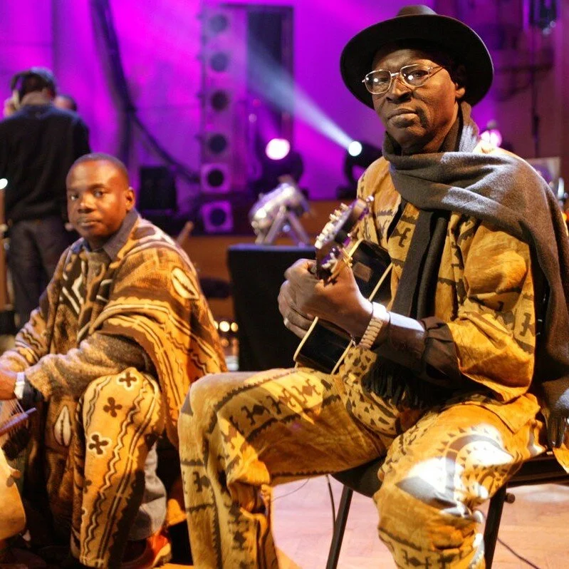 Two musicians dressed in traditional African attire performing on stage, one holding a guitar, with stage lights and equipment in the background.