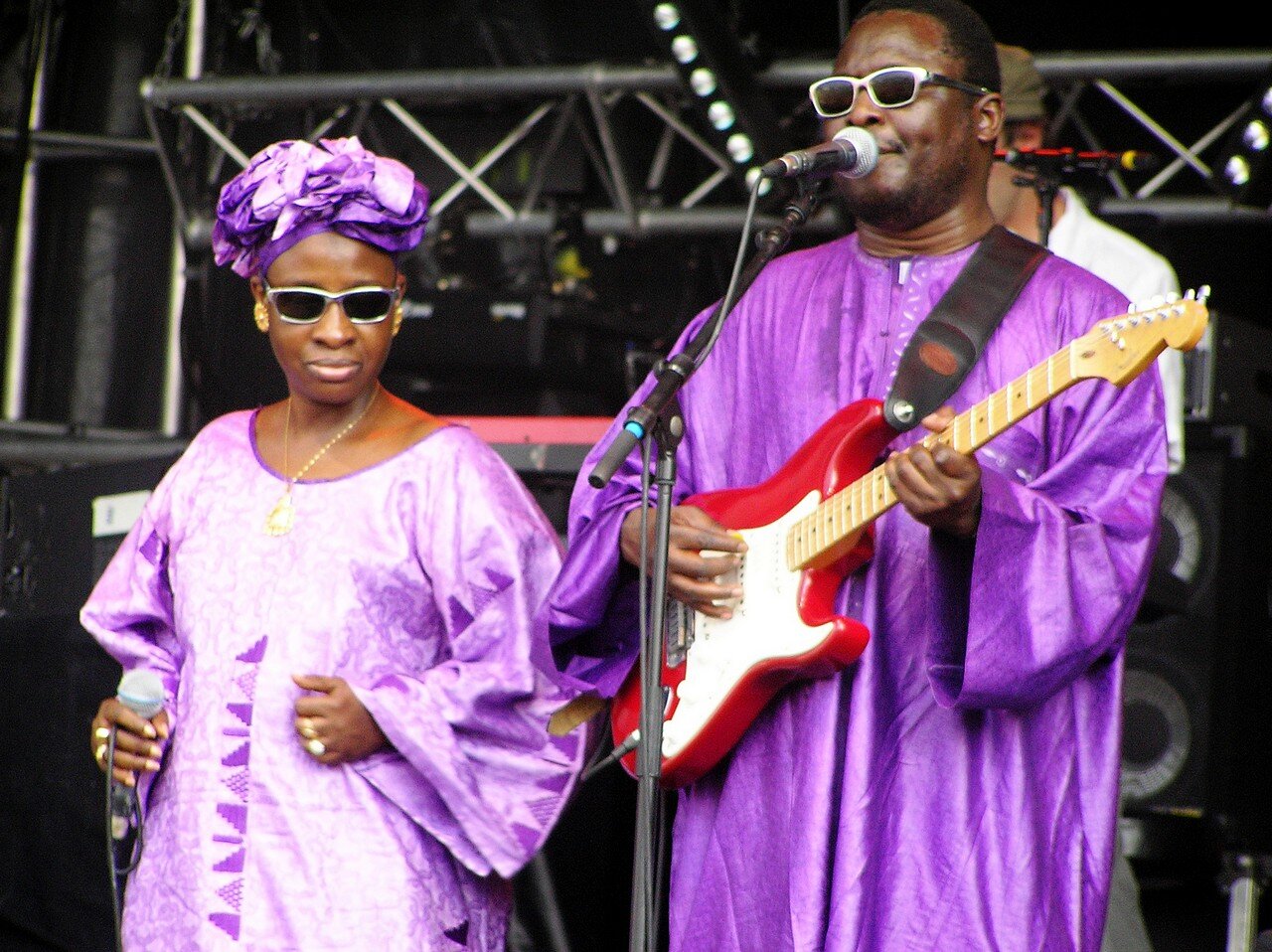 Two musicians on stage, both wearing matching purple outfits and sunglasses. The man on the right is playing an electric guitar and singing into a microphone, while the woman on the left holds a microphone and stands beside him.