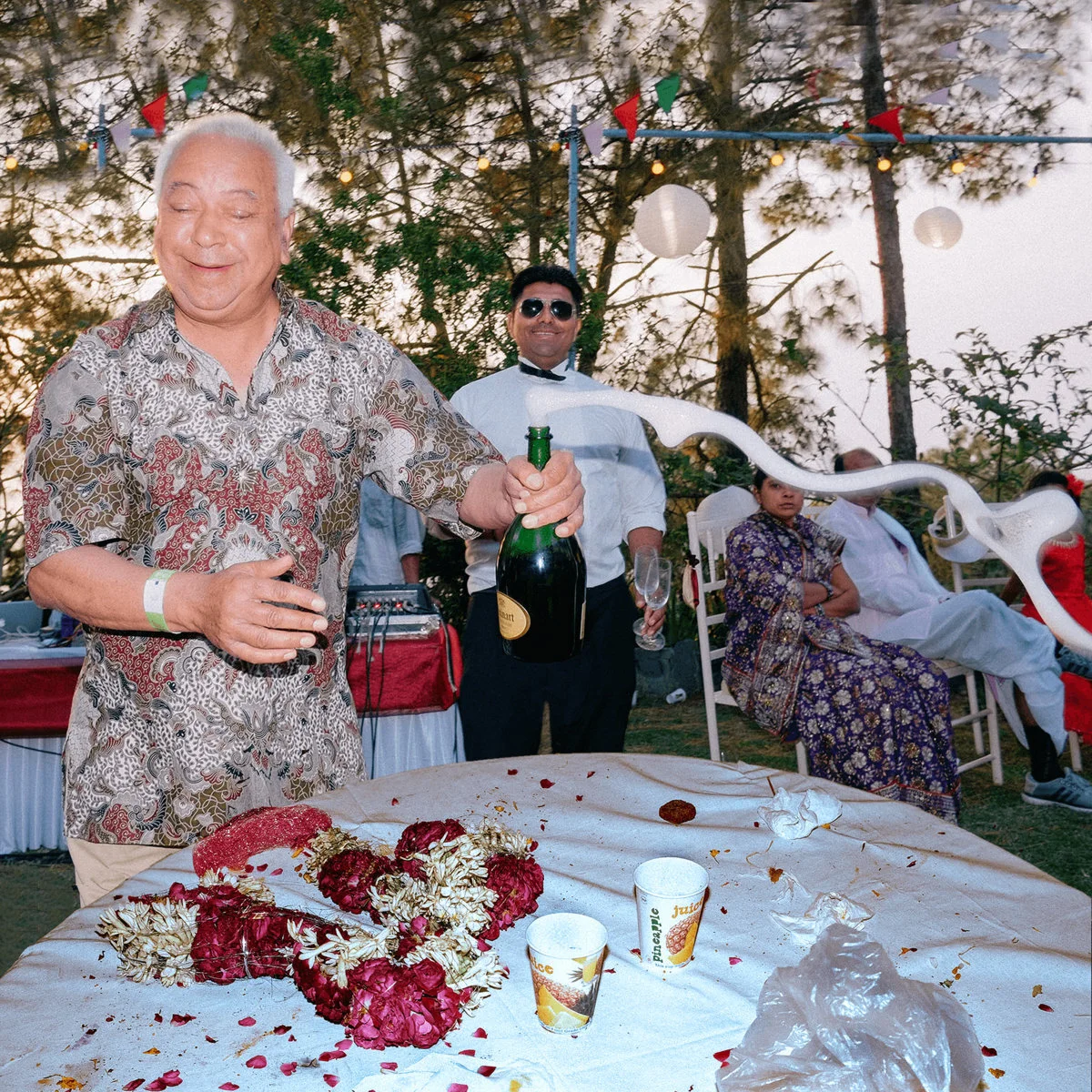 A man in a patterned shirt enthusiastically opens a champagne bottle, causing it to fizz. Behind him, a man in sunglasses stands with a champagne flute. A table in front is scattered with red and white flowers and paper cups. The scene is outdoors wi