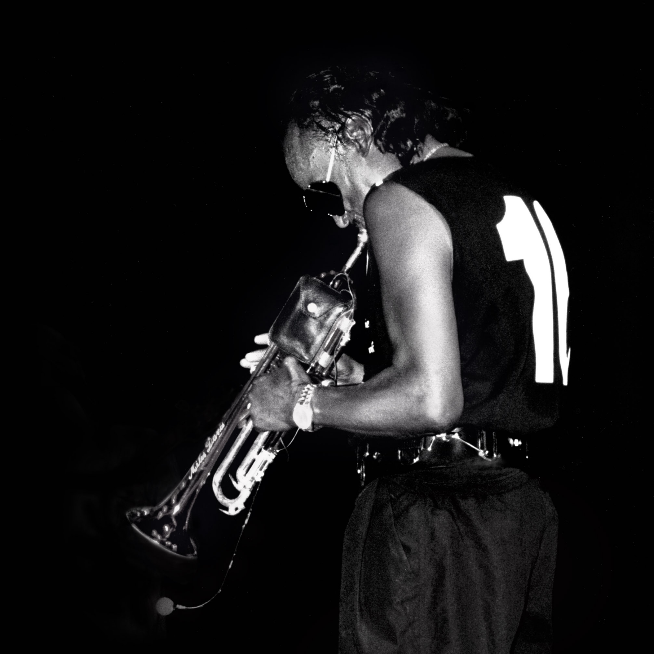 Black and white photo of a jazz musician playing a trumpet on stage, wearing a sleeveless shirt with the number 11 and sunglasses, against a dark background.
