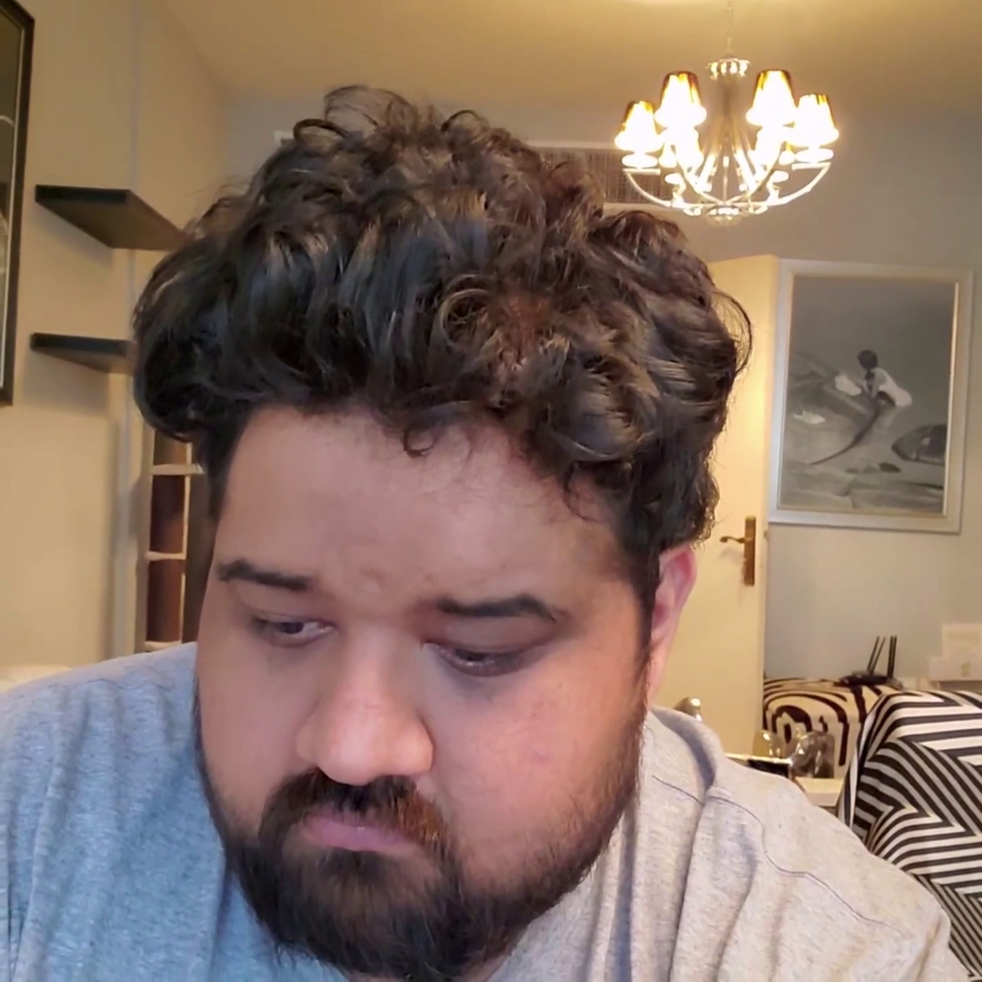 Man with curly hair and beard looking down, sitting in a room with a chandelier and artwork on the wall.