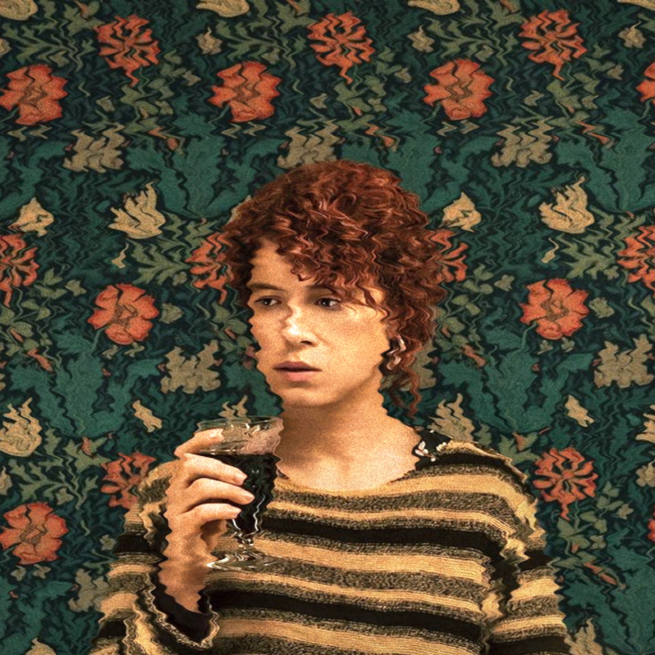 Person with curly hair holding a glass in front of a floral patterned background, wearing a striped sweater.