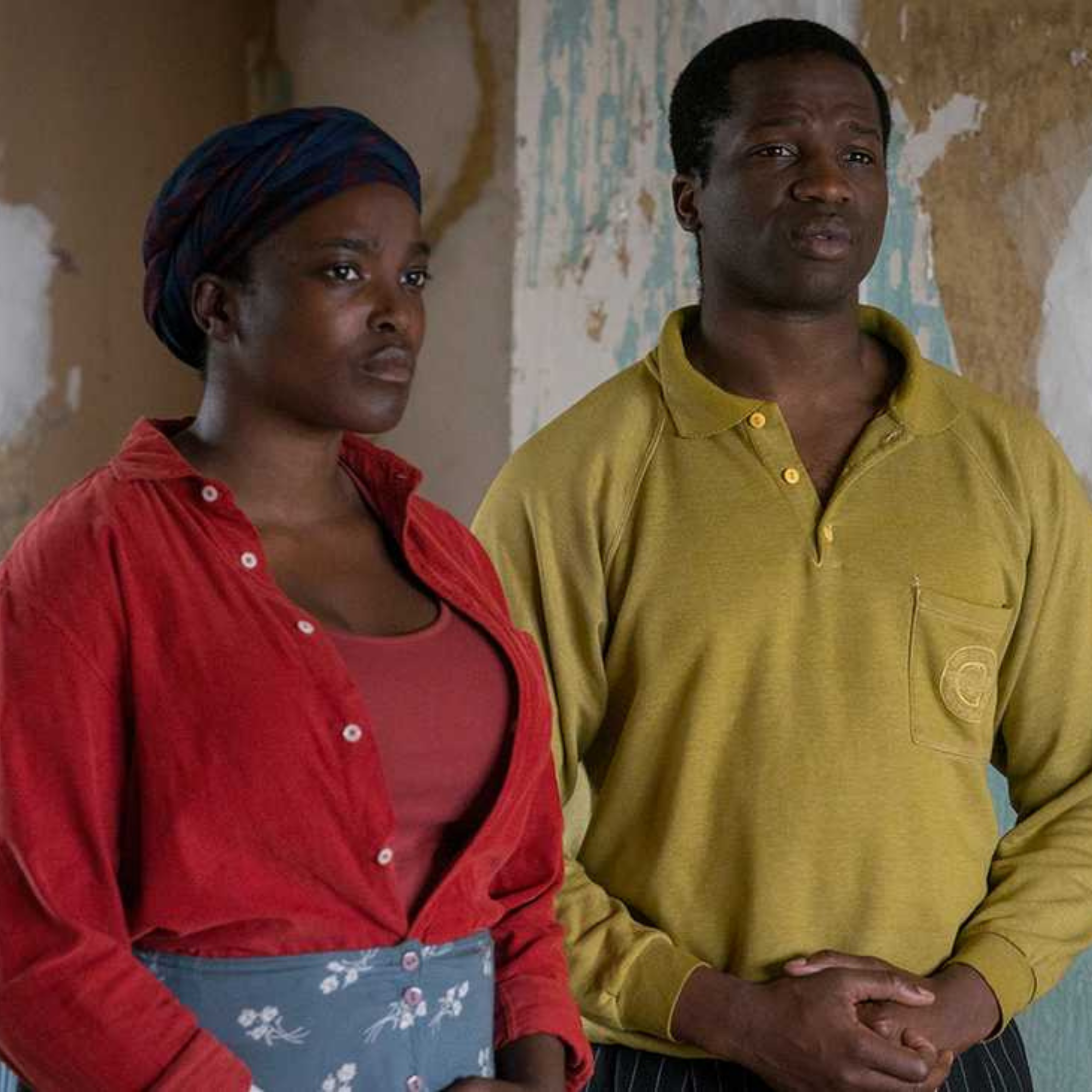 Two people standing indoors, one wearing a red shirt and headscarf, the other in a yellow long-sleeve shirt, in front of a textured wall.