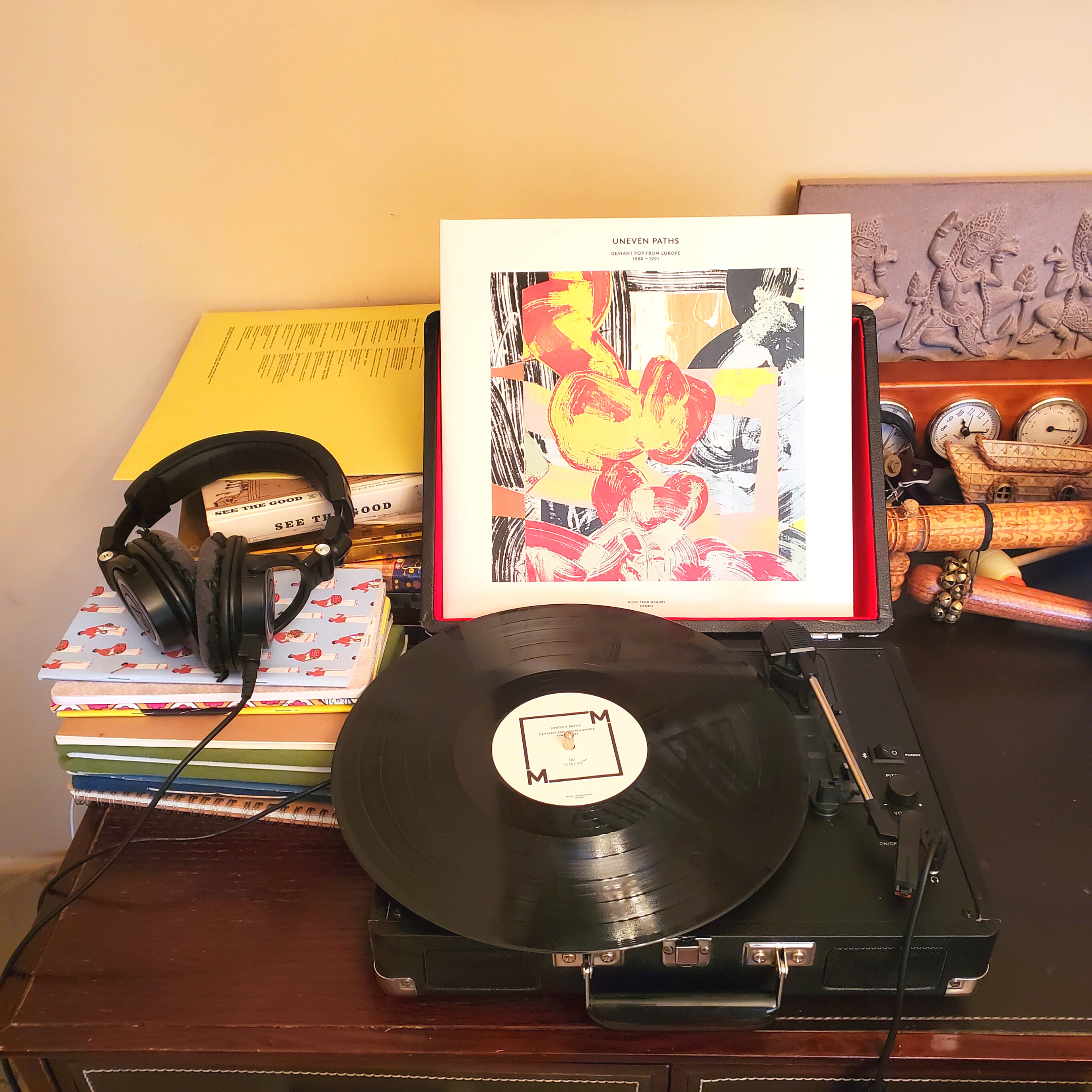 Vinyl record player with a spinning black vinyl record and an album cover on display. Headphones rest on a stack of books next to the player. Various decorative items are in the background.