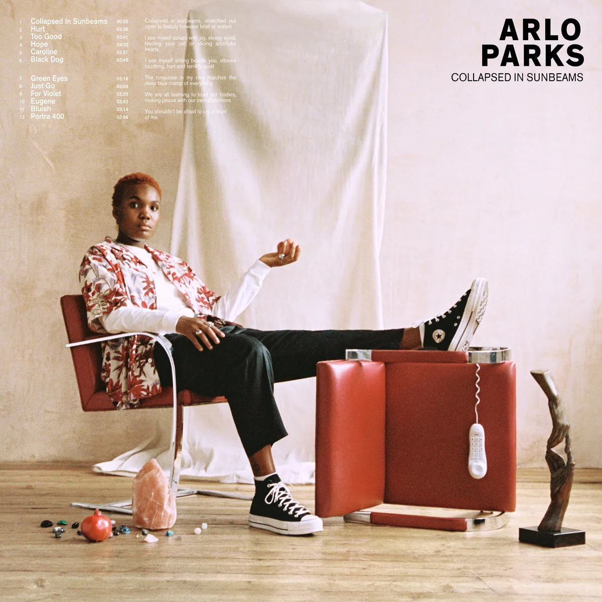 Album cover of 'Collapsed in Sunbeams' by Arlo Parks featuring an individual sitting on a red chair, wearing a floral shirt, black pants, and sneakers. They are surrounded by small objects like a salt lamp, a pomegranate, and various stones. Text lis