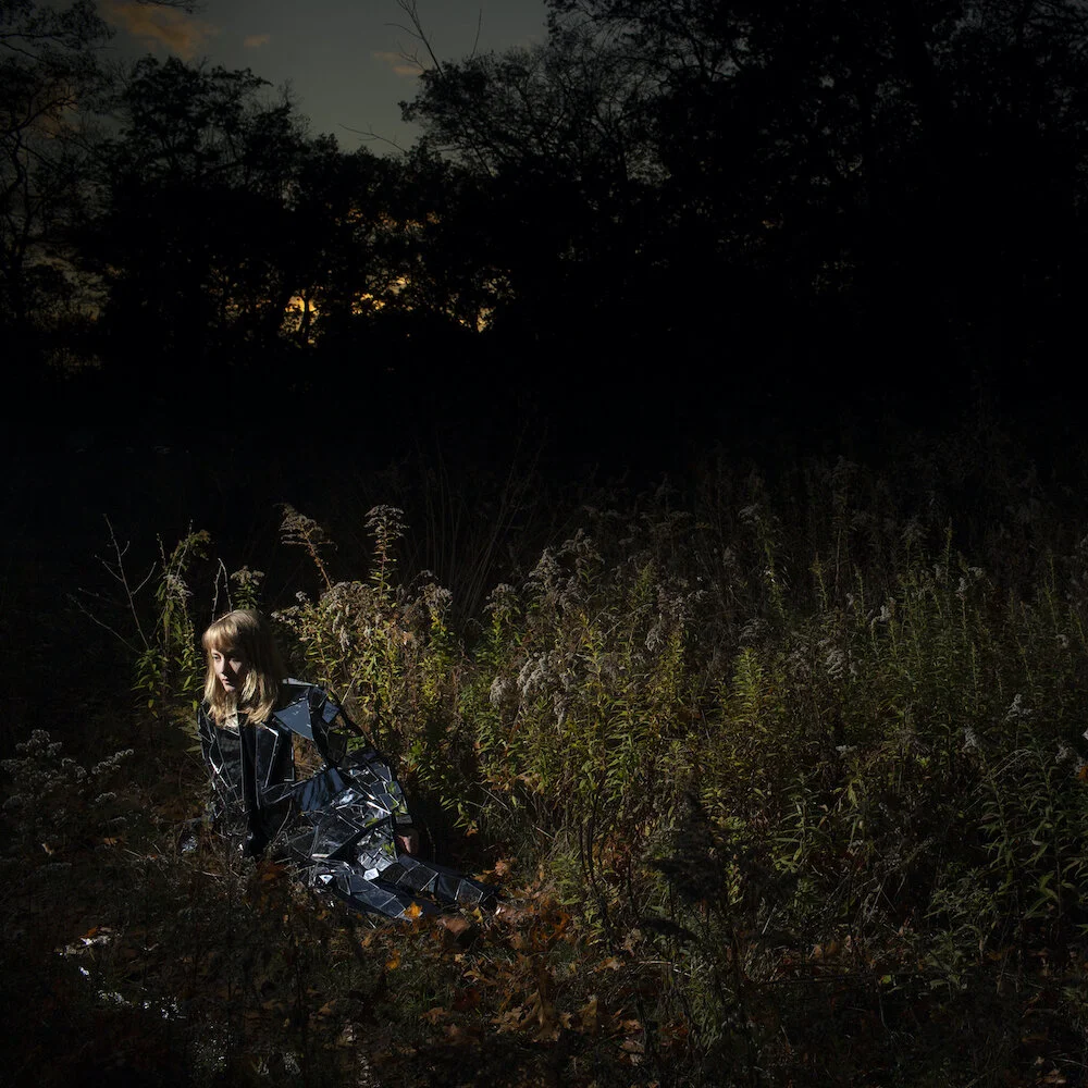Person in a reflective outfit sitting in a dark field with wild plants, against a backdrop of a silhouetted forest at dusk.