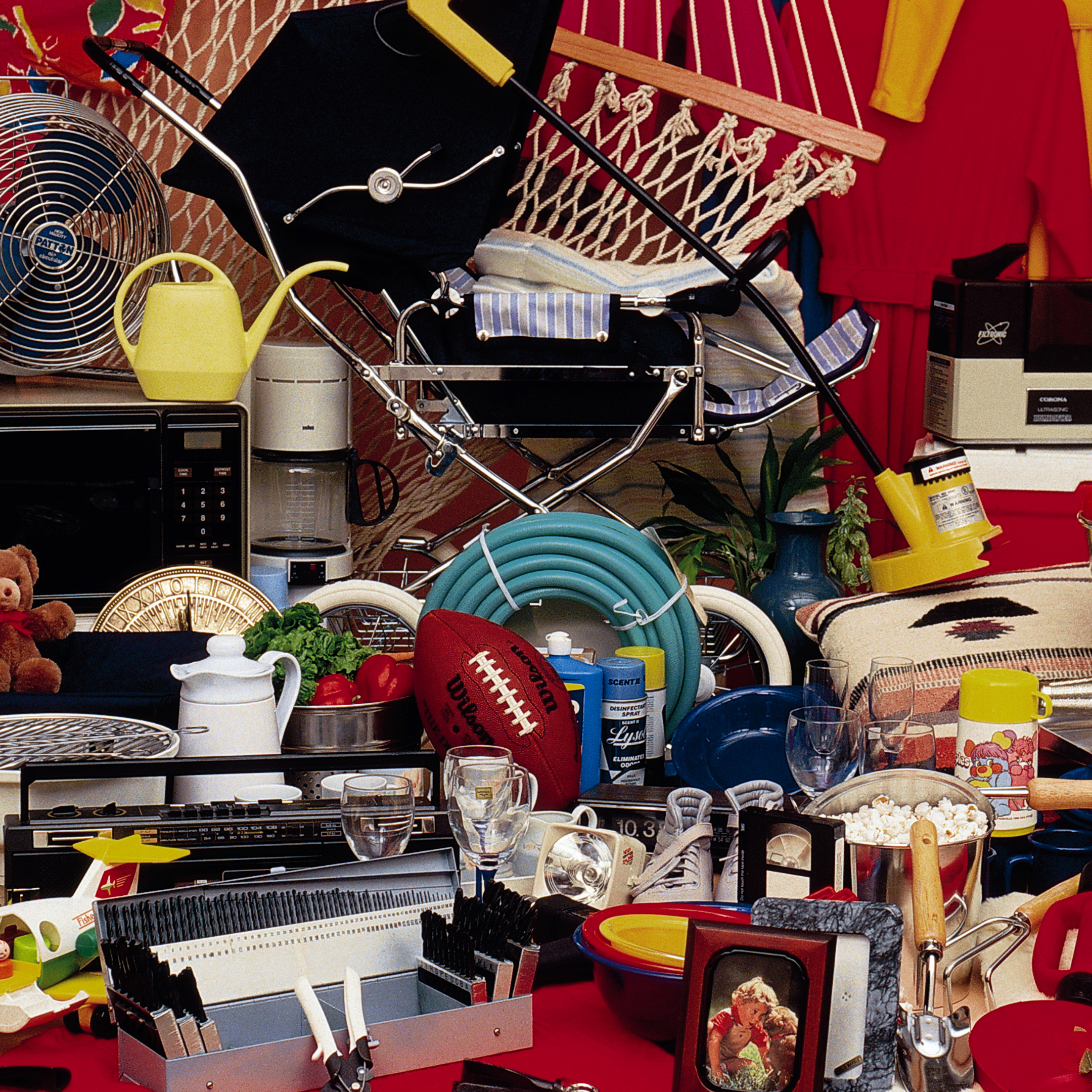 A cluttered collection of household items, including a stroller, hammock, fan, microwave, watering can, football, garden hose, cleaning products, and kitchen utensils. Various decorations, cookware, and beverages are also visible.
