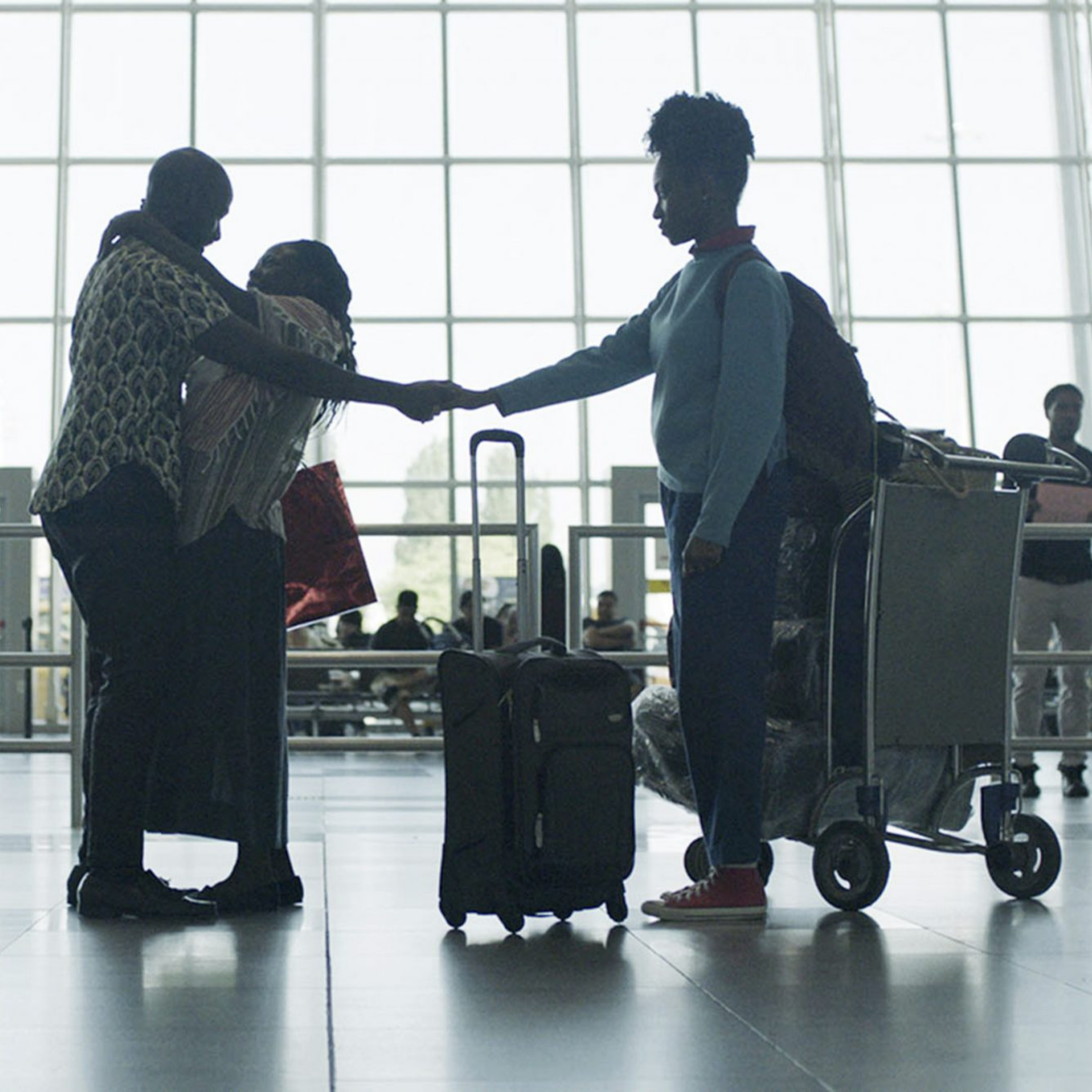 Three people saying goodbye in an airport, one with a suitcase, another with a luggage cart, and a window in the background.