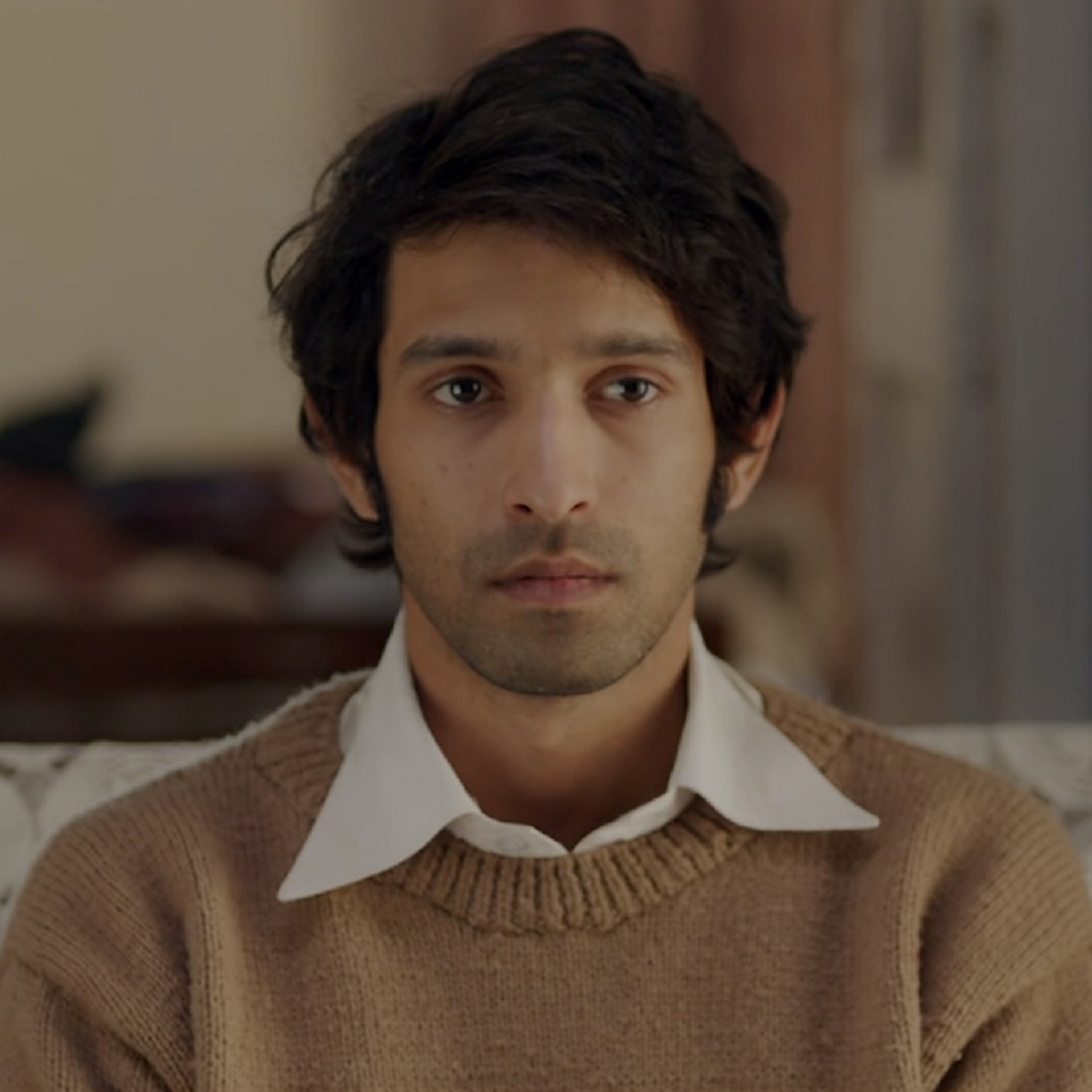 A man wearing a brown sweater and white collared shirt sitting indoors.