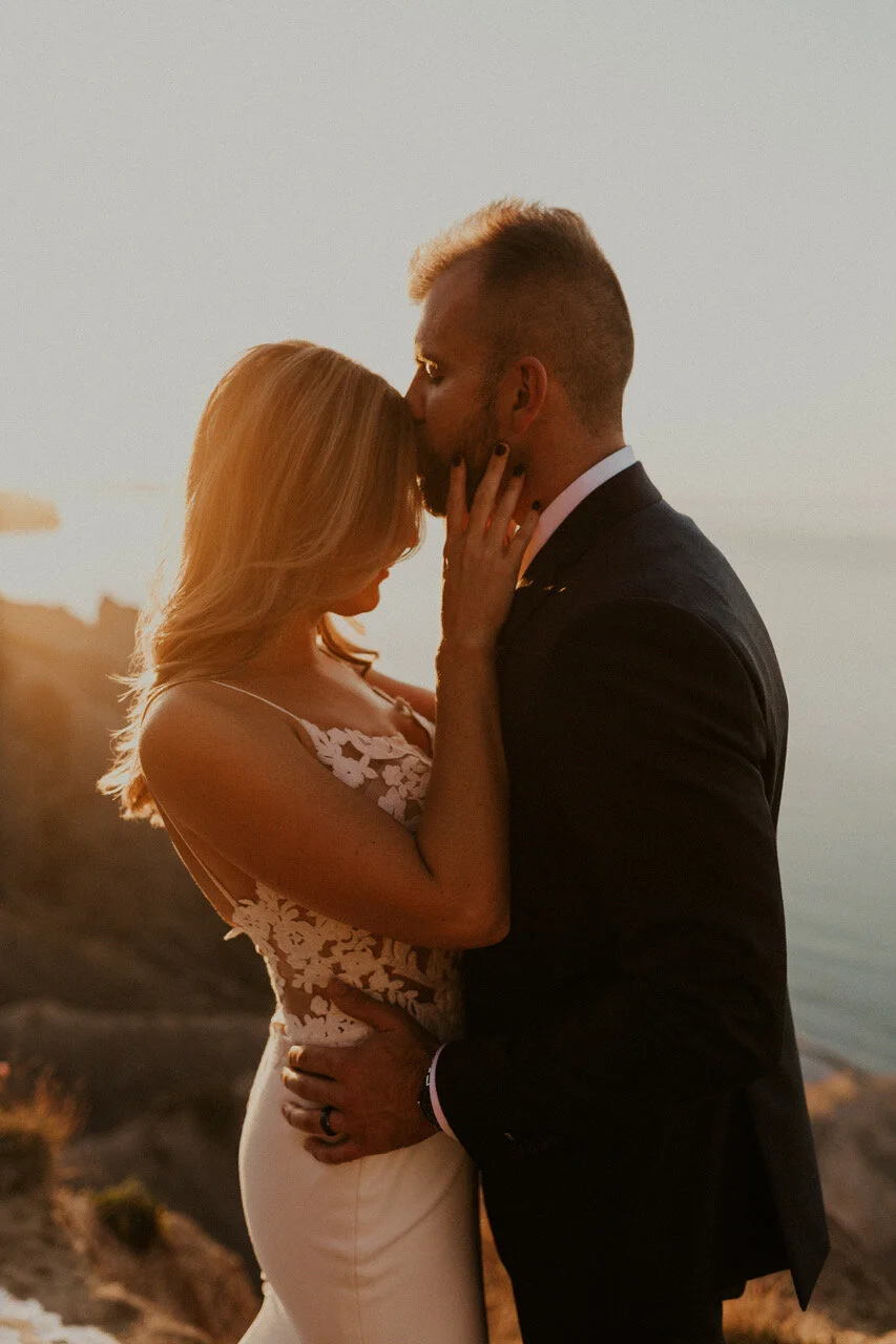 A couple shares a kiss at sunset on a rocky coastline, with soft lighting and a calm sea in the background.