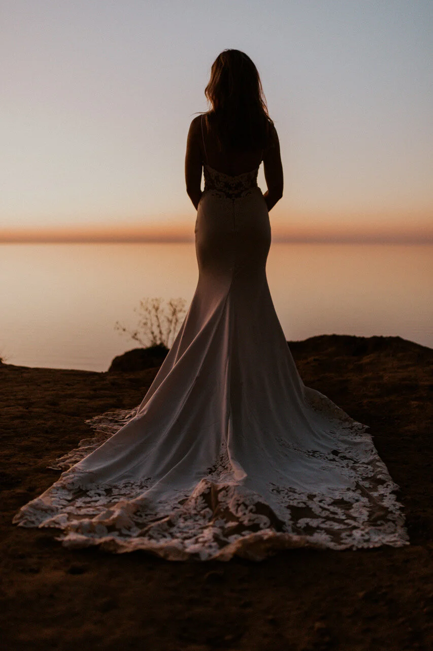 A woman in a long wedding dress standing outdoors on a rocky surface at sunset, facing a body of water.