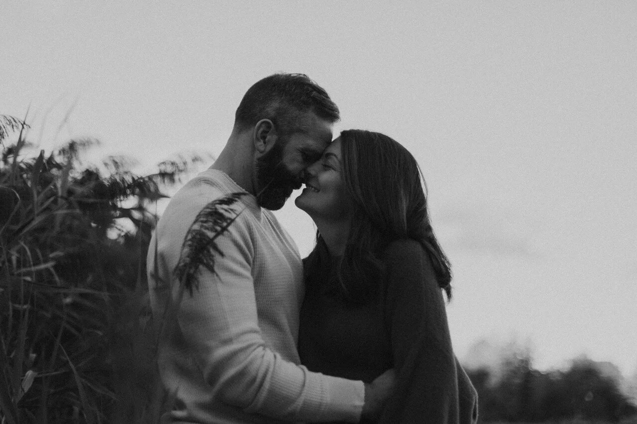 A black and white photo of a couple standing close with foreheads touching and smiling in an outdoor setting.