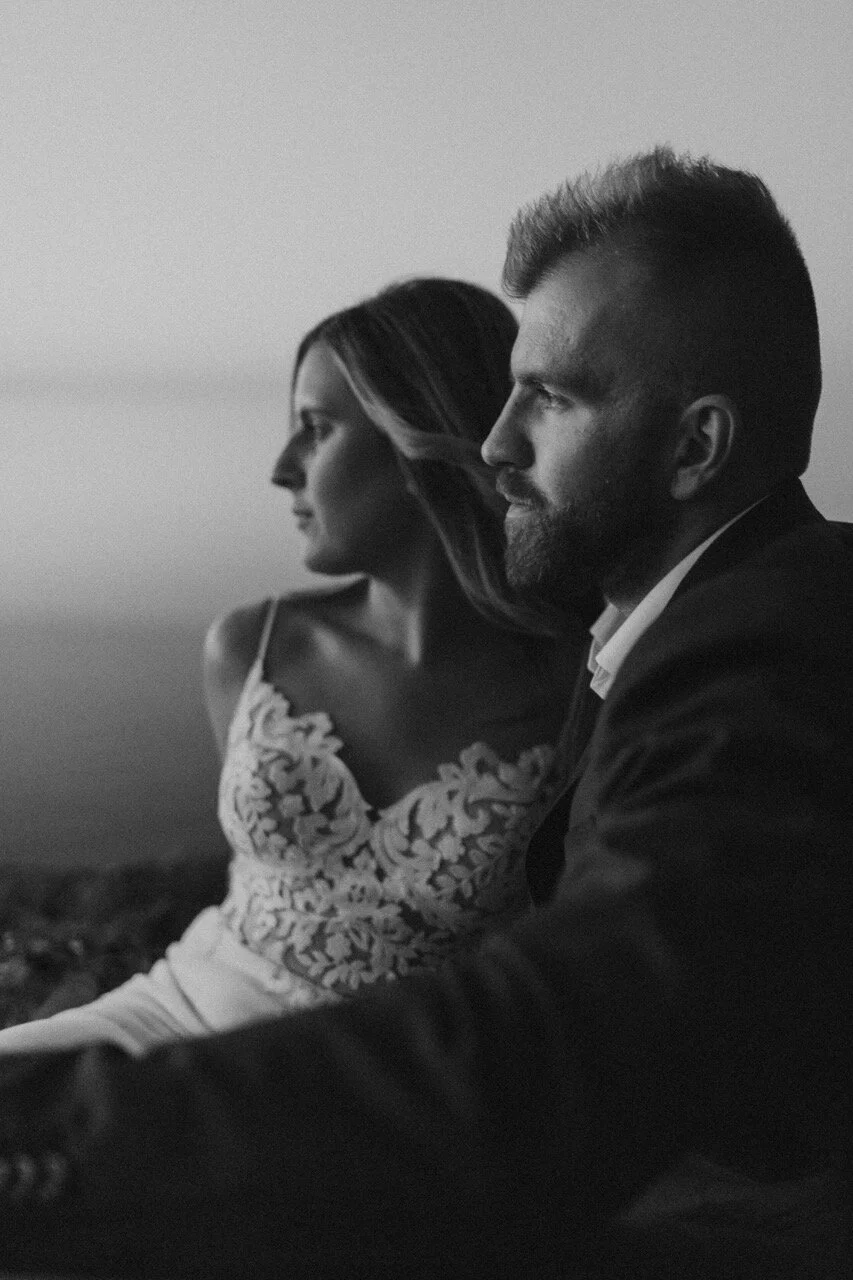 A black-and-white photo of a man and woman sitting side by side, facing to the right. The woman is wearing a dress with floral embroidery, and the man is dressed in a suit. They are both looking into the distance with a calm and contemplative express