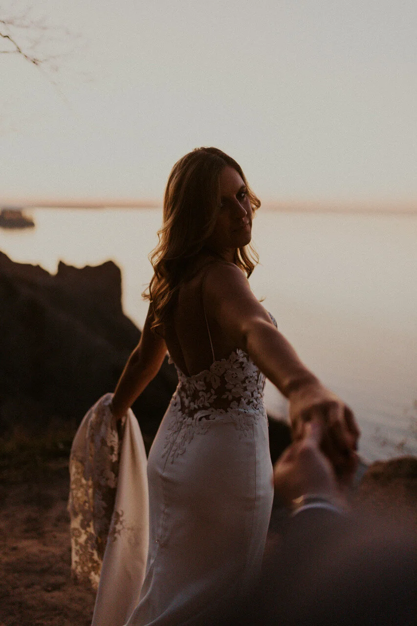 A woman in a wedding dress holding someone else's hand near a lakeshore at sunset.