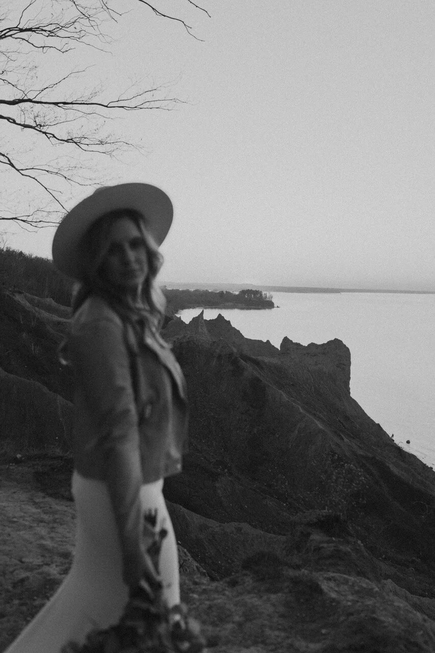 A woman wearing a wide-brimmed hat and a jacket stands on a rocky cliff overlooking a body of water with a distant shoreline and leafless trees.