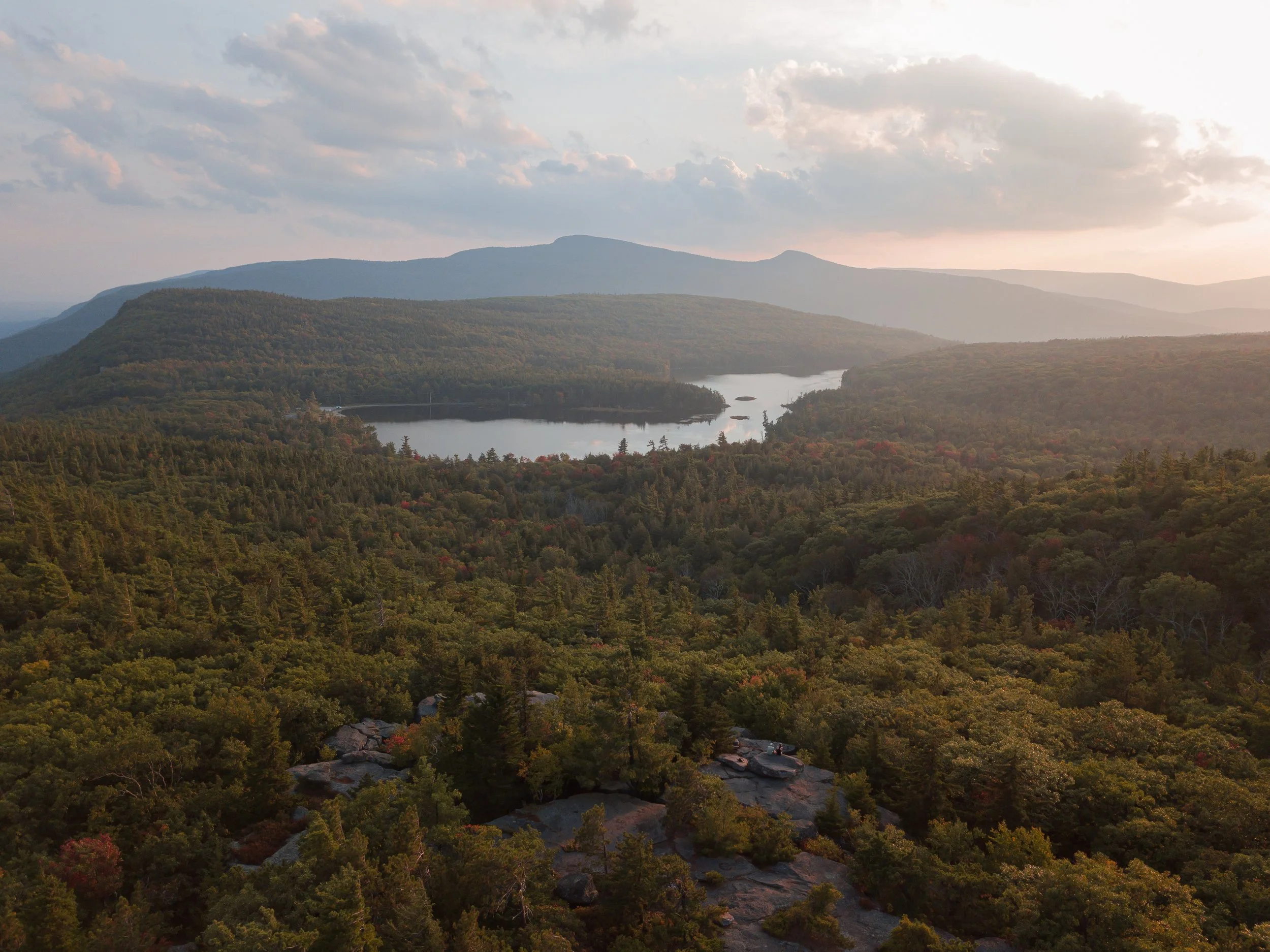 A panoramic view of a mountainous landscape with a forested area, a body of water, and distant mountain ranges under a partly cloudy sky.