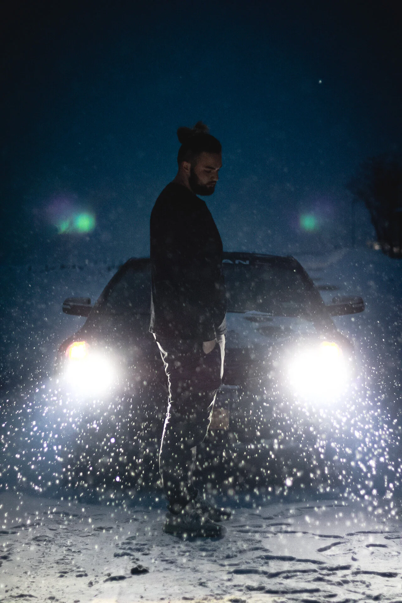 Man at night standing in snowy landscape with snow falling, backlit by car headlights, wearing dark clothing, with a man bun, looking down.