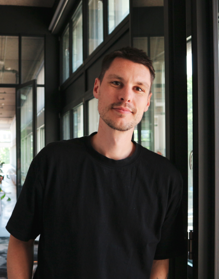 Een jonge man met kort donker haar en een lichte huid staat binnen bij grote ramen, gekleed in een zwart T-shirt, en kijkt met een lichte glimlach naar de camera.