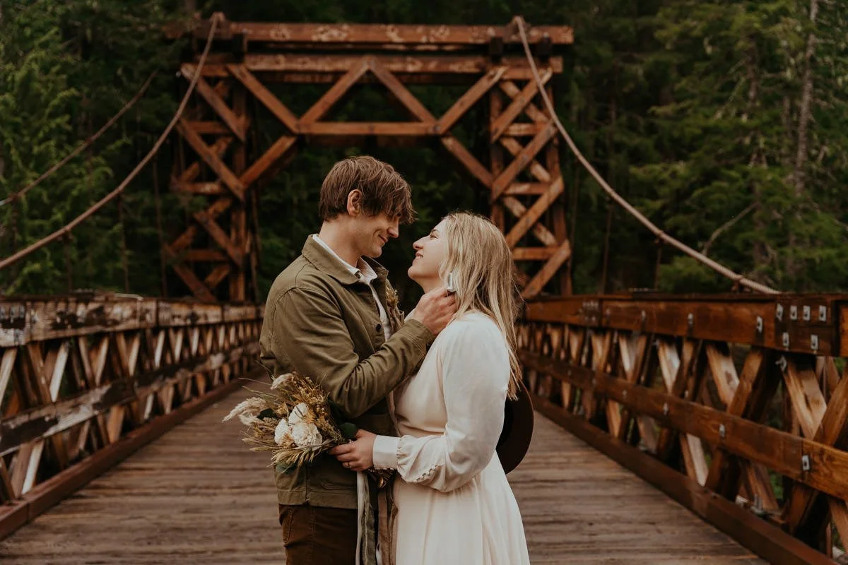 A couple stands close together on a wooden bridge surrounded by trees, gazing into each other's eyes. The man holds a small bouquet of dried flowers, and the woman wears a white dress and a black hat.