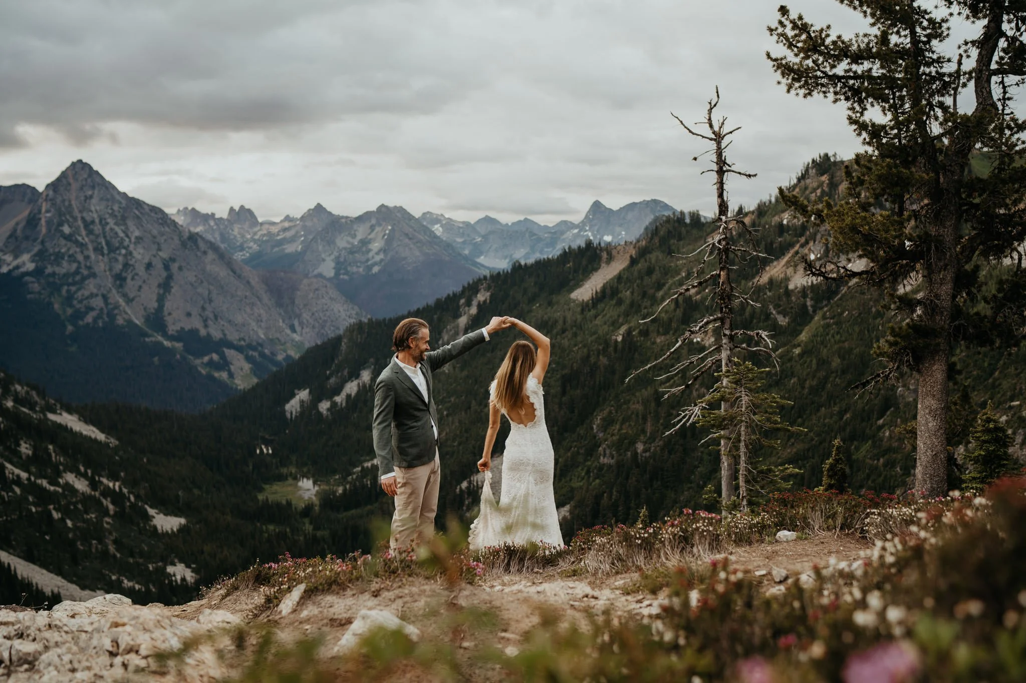 A man in a gray blazer and a woman in a white dress dancing on a mountain overlook with a mountain range in the background.