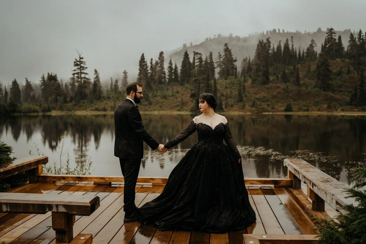 A couple in formal black wedding attire holding hands on a wooden platform by a lake with a moody, mountainous forested background.