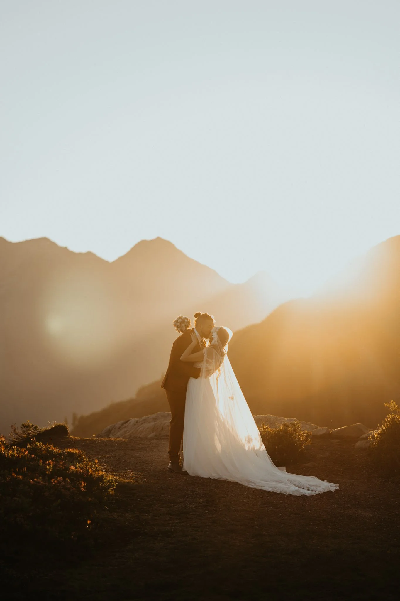 A couple in wedding attire sharing a kiss at sunset in a mountainous landscape.