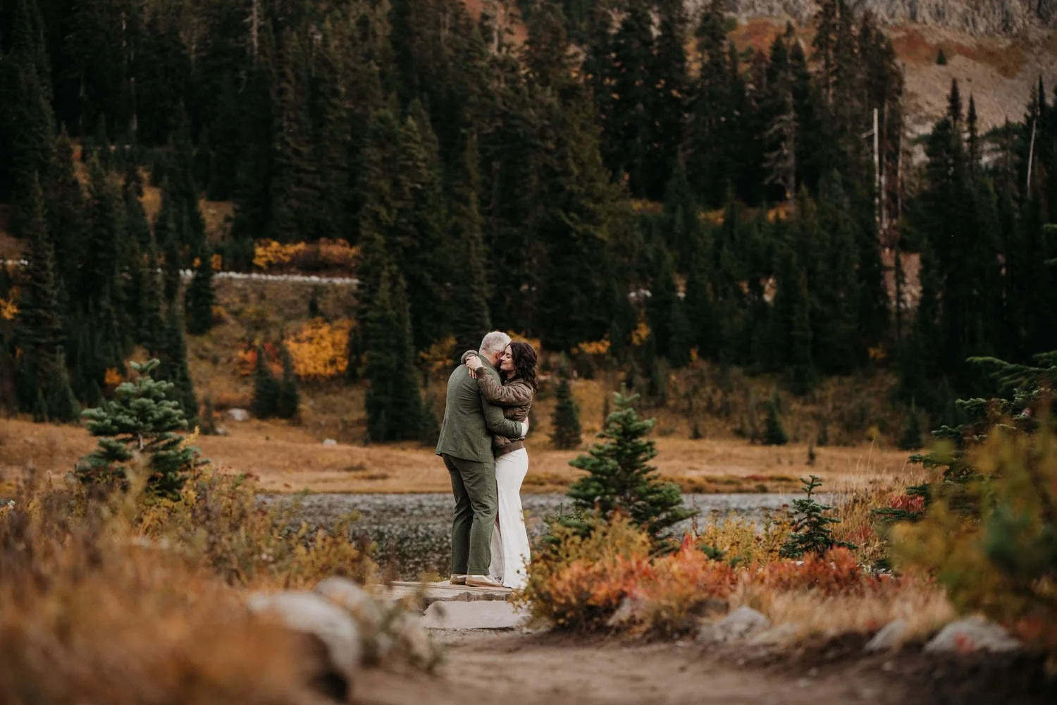 A couple hugging on a rocky path in a forested area with autumn foliage and mountains in the background.
