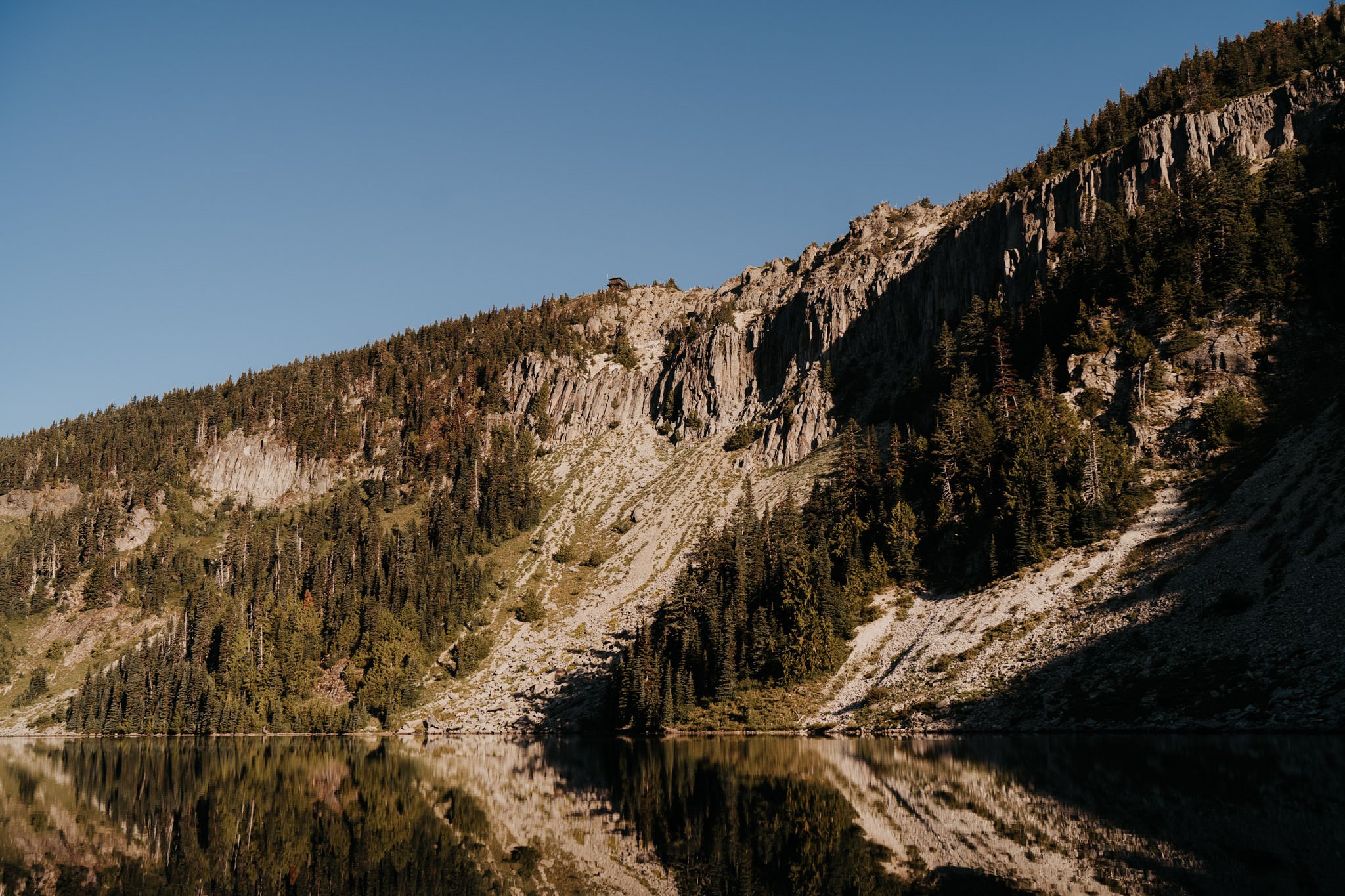 Reflection of a tree-covered mountain with rocky cliffs in a calm lake under a clear blue sky.