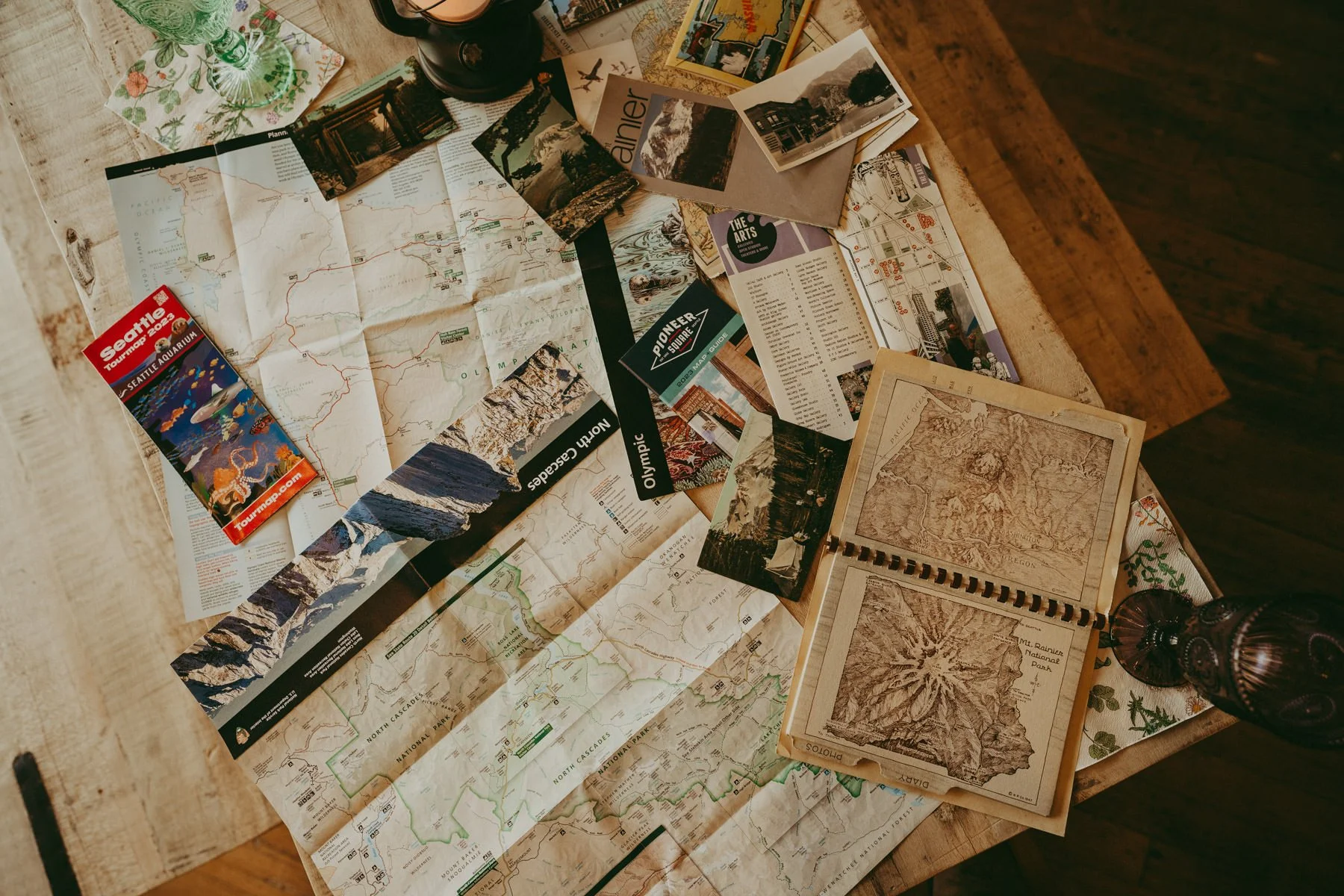 Spread of various maps, brochures, postcards, and travel guides on a wooden table, including a large folded map, a Seattle Aquarium brochure, and vintage-style paper maps.
