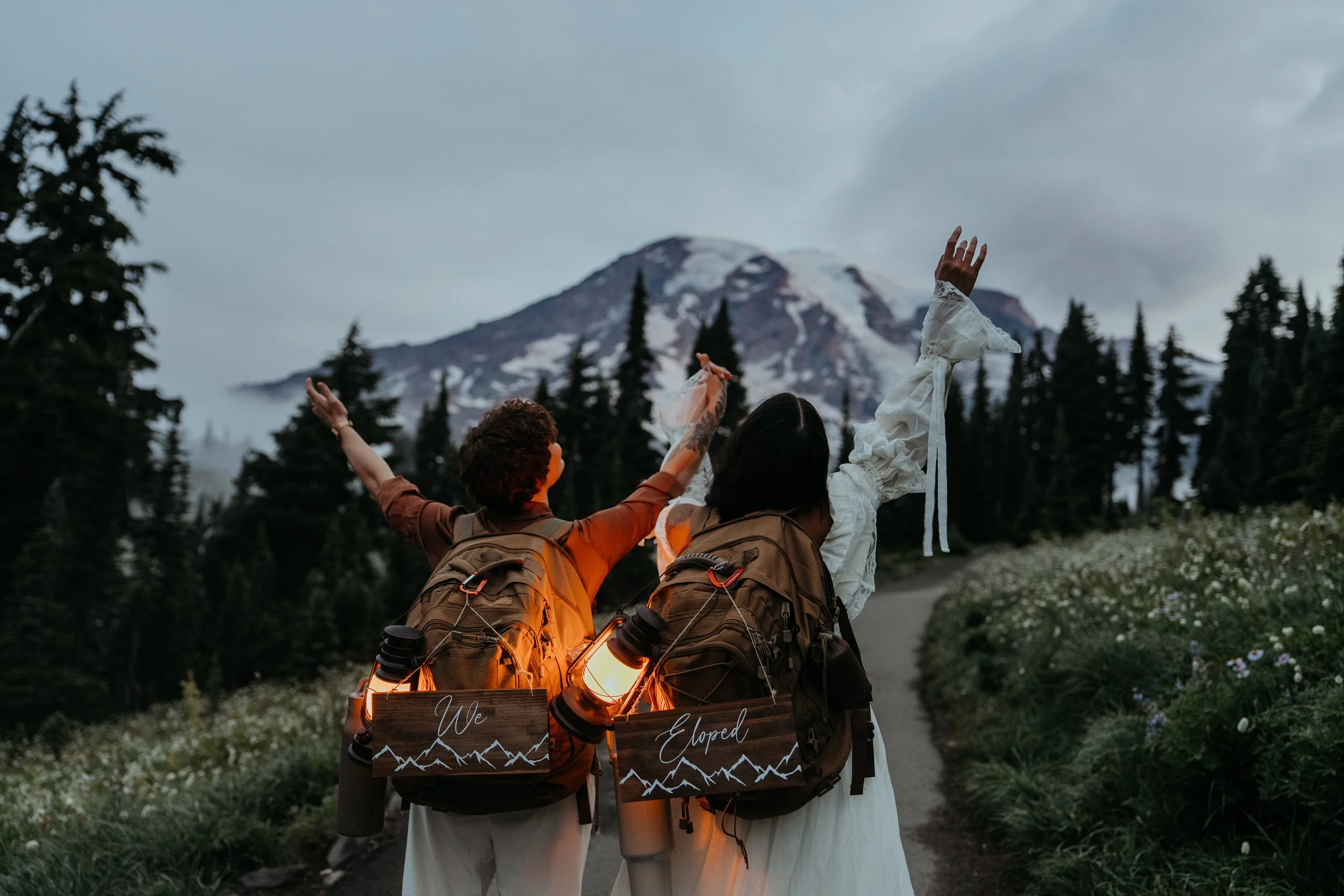 Two hikers with backpacks and lanterns, one with a sign saying "We" and the other with "Elope" on their backpacks, celebrating in front of a mountain with snow and evergreen trees during dusk.