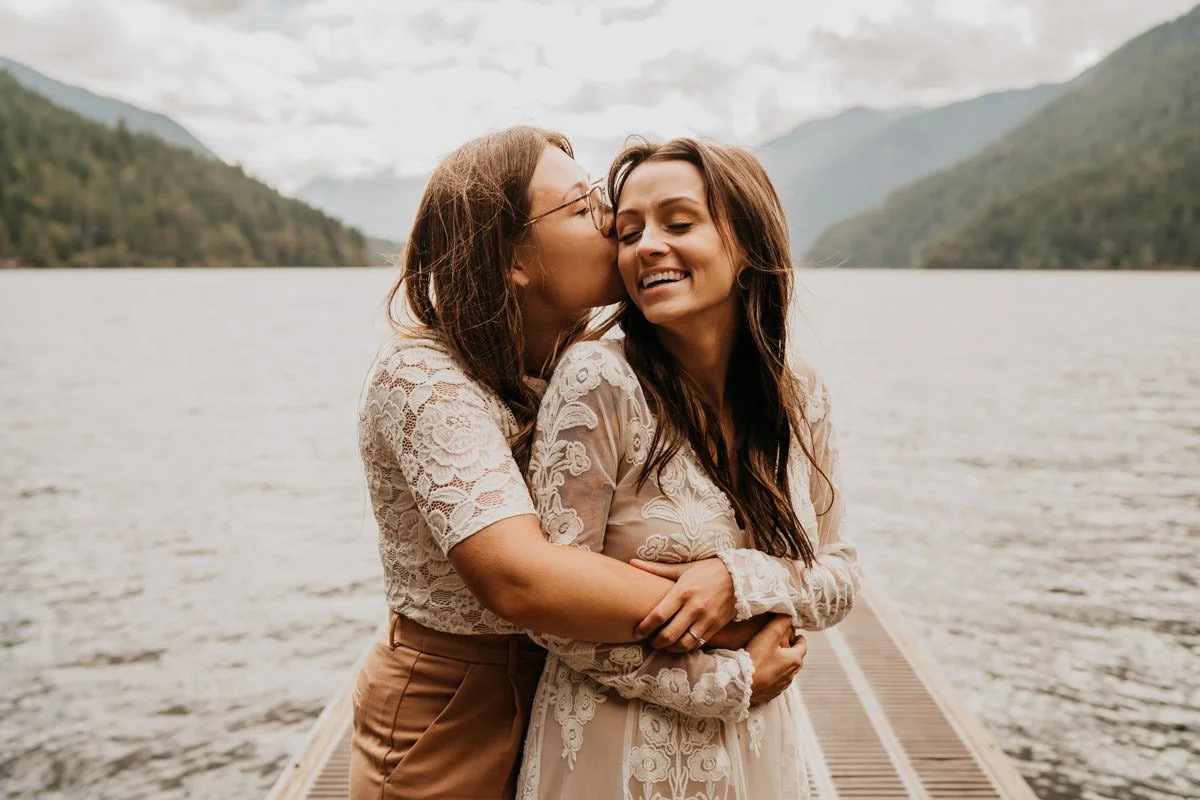 Two women embrace on a wooden dock by a lake surrounded by mountains, with one kissing the other's cheek and both smiling.