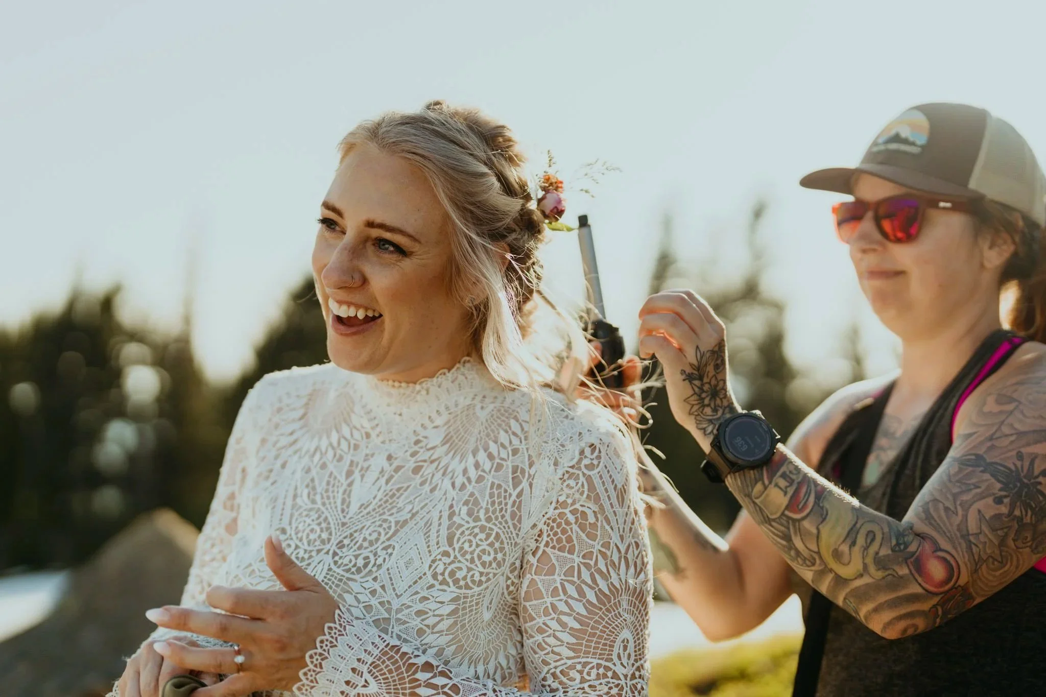 A smiling bride with a floral hairpiece is getting her hair styled outdoors by a tattooed woman wearing sunglasses and a cap during late afternoon sunlight.