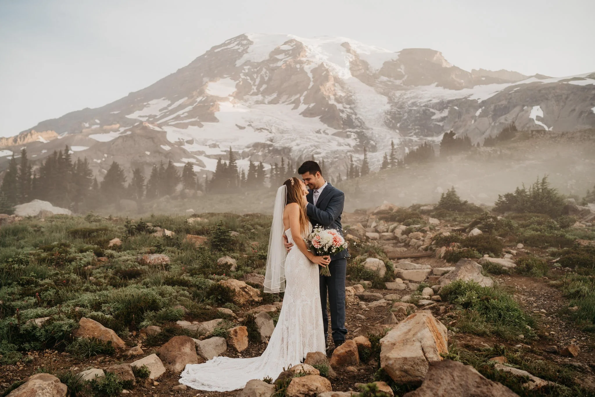 A bride and groom standing closely on a mountain trail, with the bride holding a bouquet of flowers, in front of a large snow-capped mountain and misty landscape.