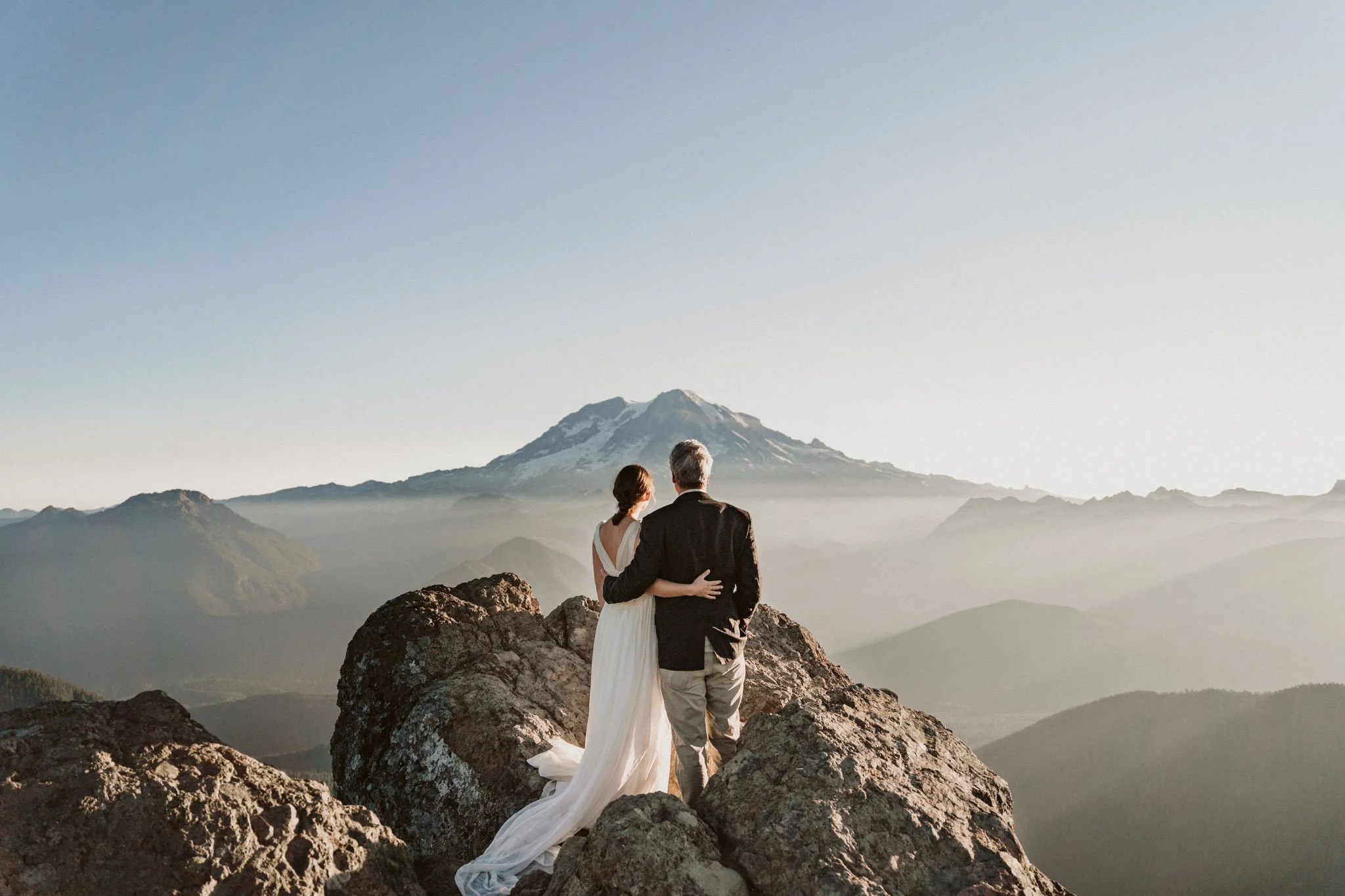 Couple in wedding attire standing on rocks on a mountain with snow-capped peaks in the background