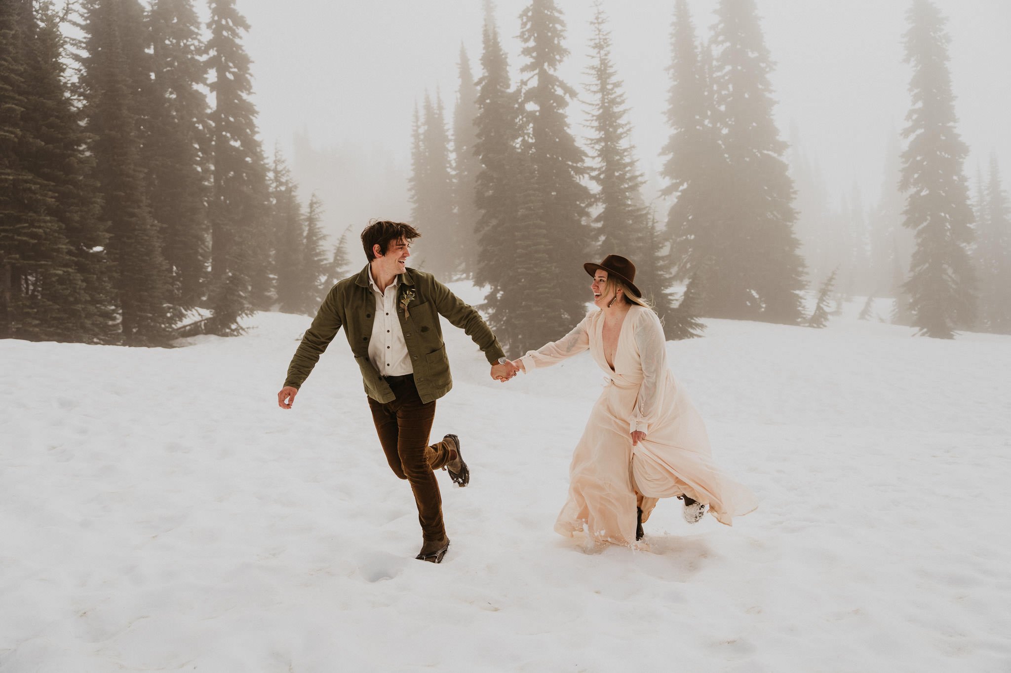 A couple holding hands and running in a snowy forest with tall pine trees, fog, and overcast sky.