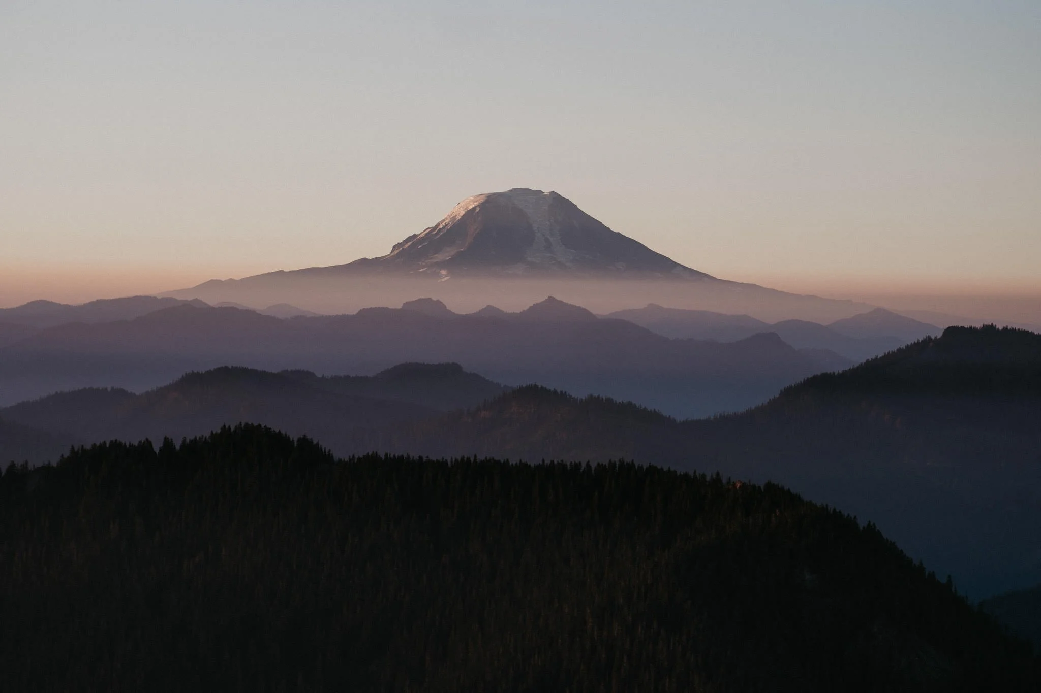 Snow-capped Mount Fuji with mountain ranges and forests in the foreground during sunset or sunrise.
