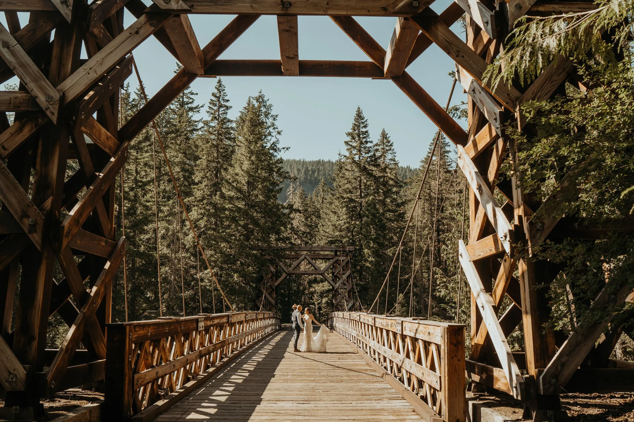A bride and groom holding hands in wedding attire standing on a wooden bridge in a forest with tall trees under a clear blue sky.