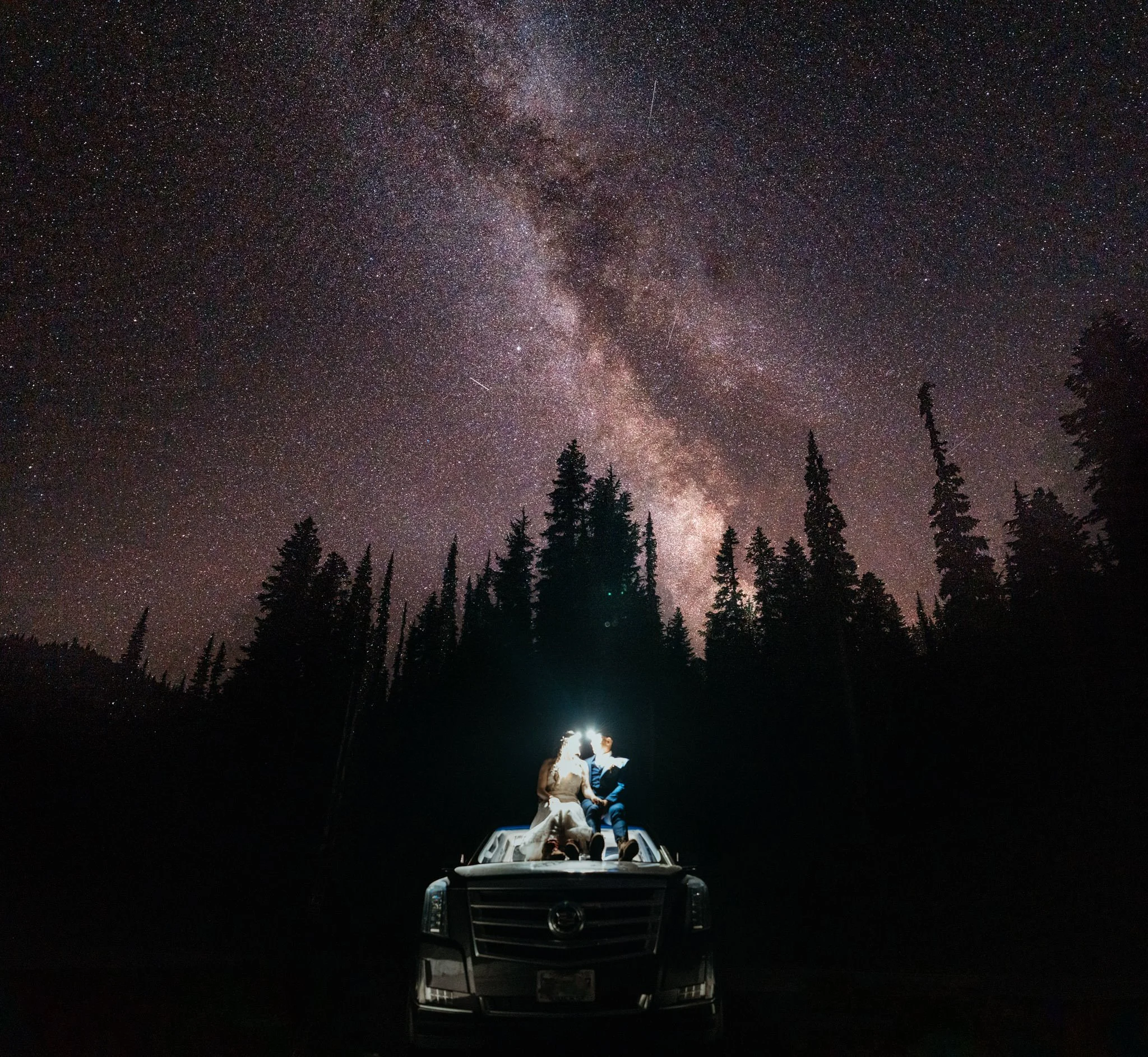 Two people sitting on the roof of a Cadillac, holding a flashlight, under a starry night sky with the Milky Way galaxy and tall pine trees in the background.