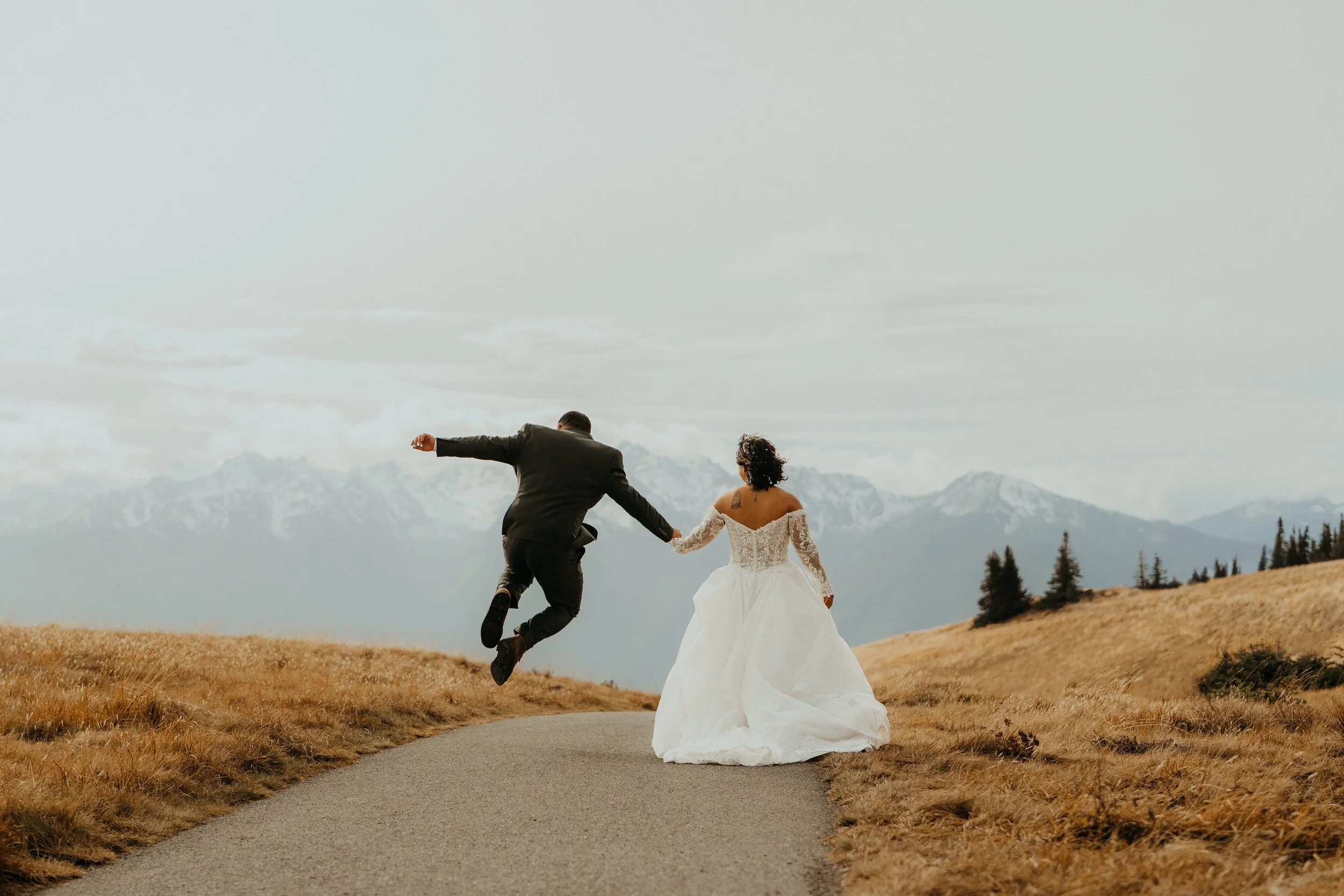 A bride and groom holding hands and dancing on a path in a mountain landscape with snow-capped peaks in the background.