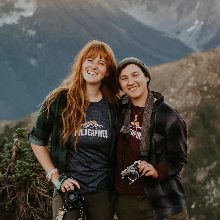 Two photographers Ashton and Tess smiling outdoors with mountain scenery in the background. Both hold cameras and wear jackets and t-shirts with 'WILDERPINES' logo.