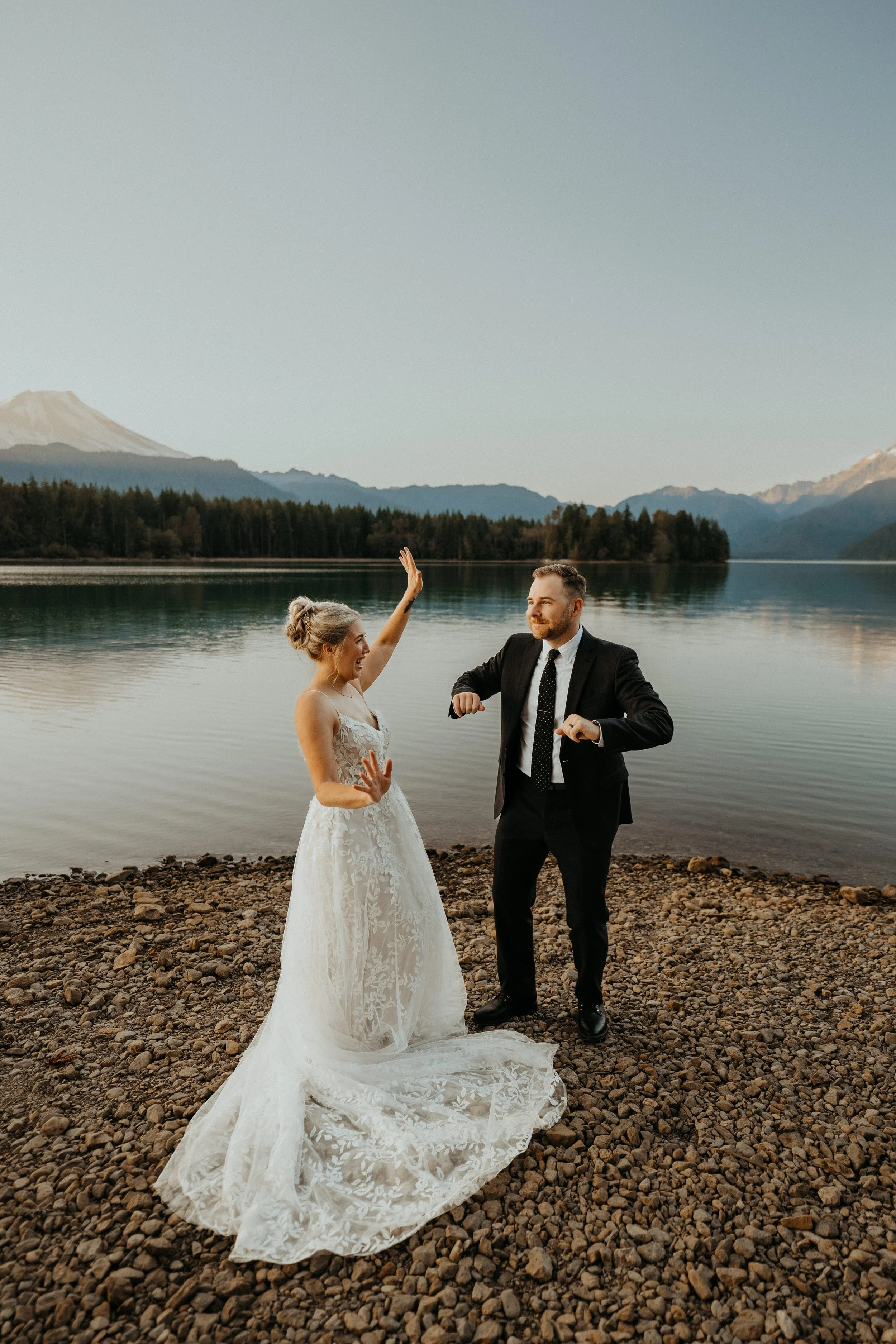 A bride in a white wedding gown and a groom in a black suit dance and celebrate on a rocky lakeshore with mountains and trees in the background.