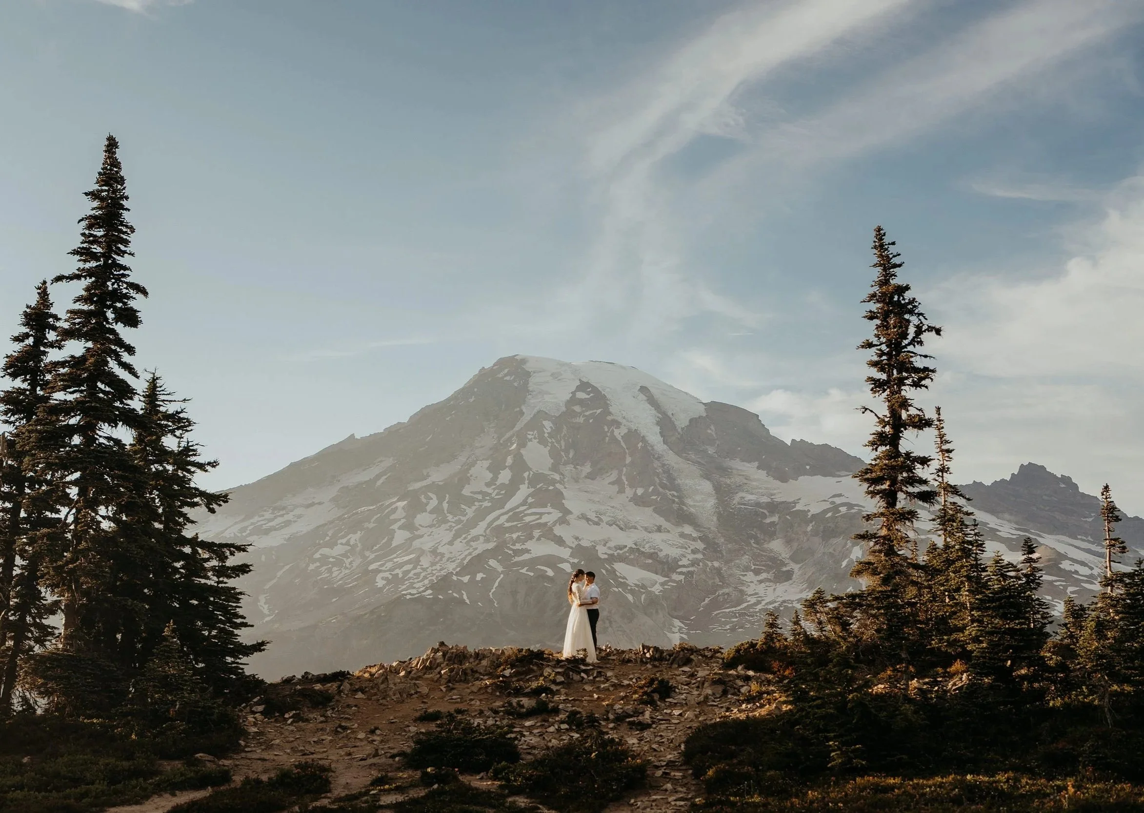 A couple in wedding attire standing on rocky terrain surrounded by tall evergreen trees, with a snow-capped mountain in the background under a partly cloudy sky.
