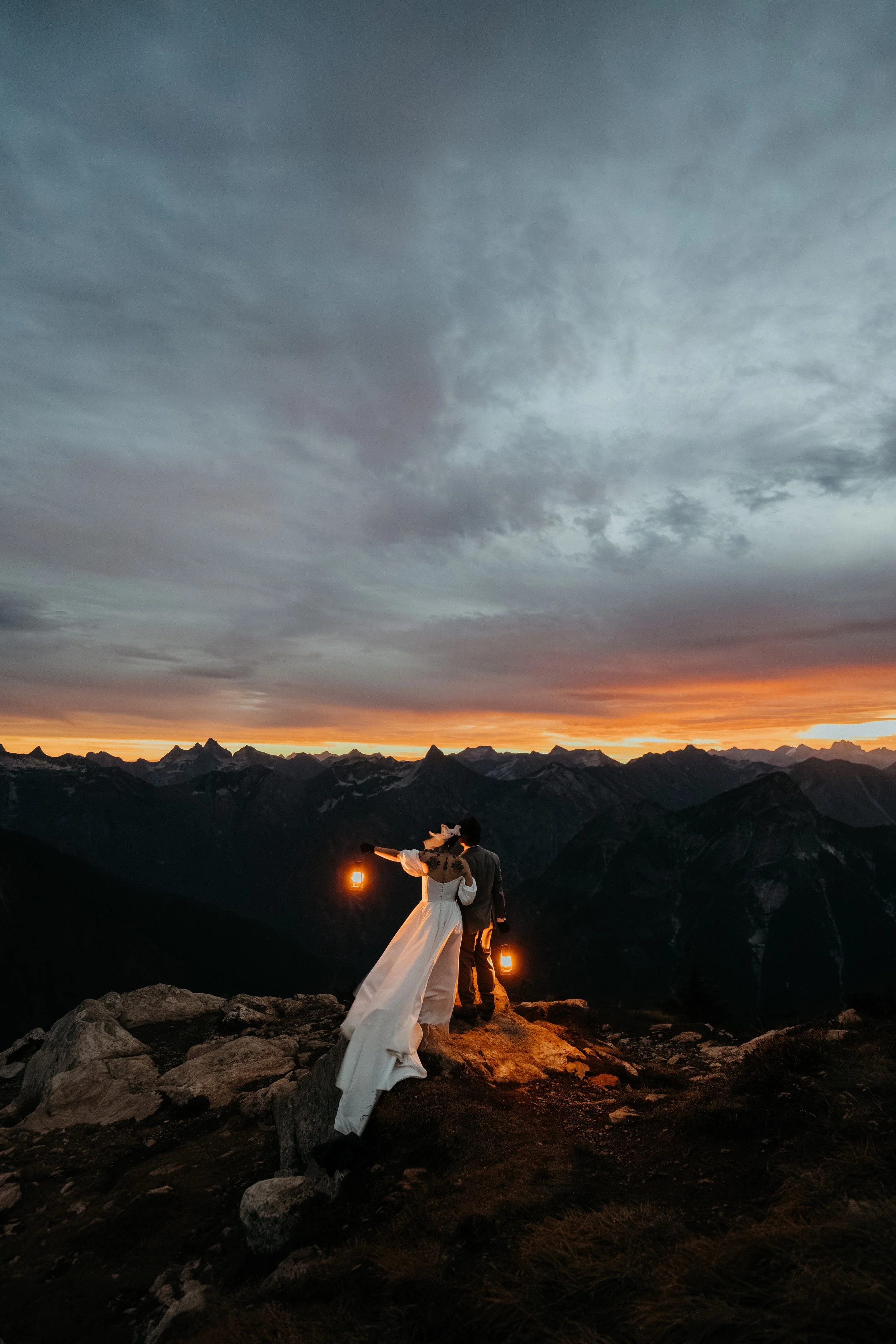 A couple in wedding attire standing on a rocky mountain ridge at sunset, holding lanterns with mountains in the background and a cloudy sky overhead.
