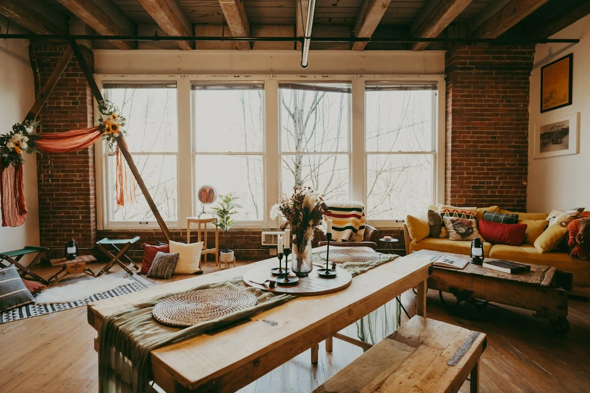 Cozy living room with large windows, exposed brick walls, wooden ceiling beams, and eclectic furniture including a yellow sofa, patterned pillows, a wooden coffee table, and a decorated dining table.