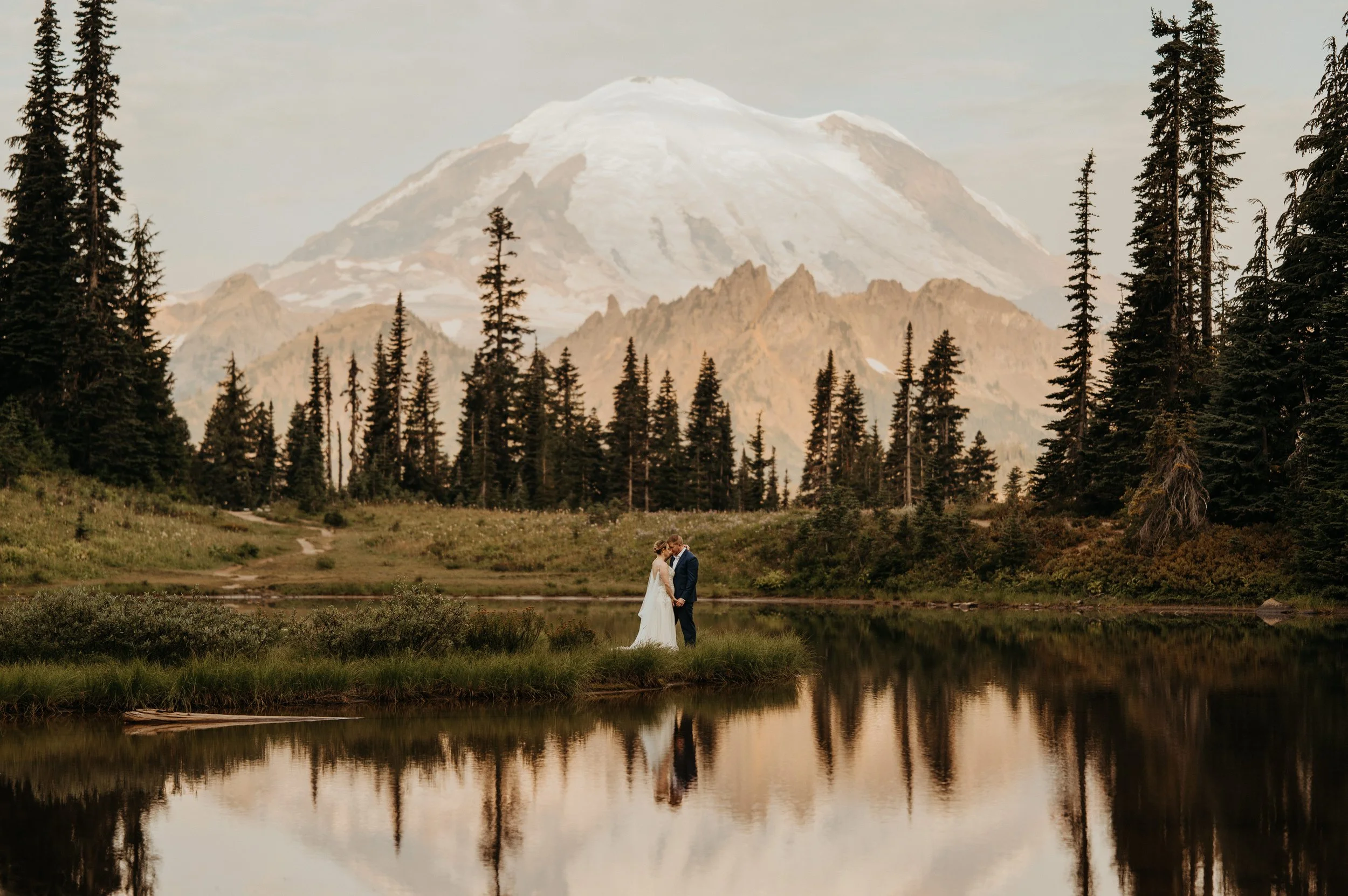 A bride and groom in wedding attire hugging by a small lake surrounded by trees and mountains, with a snow-capped mountain in the background.