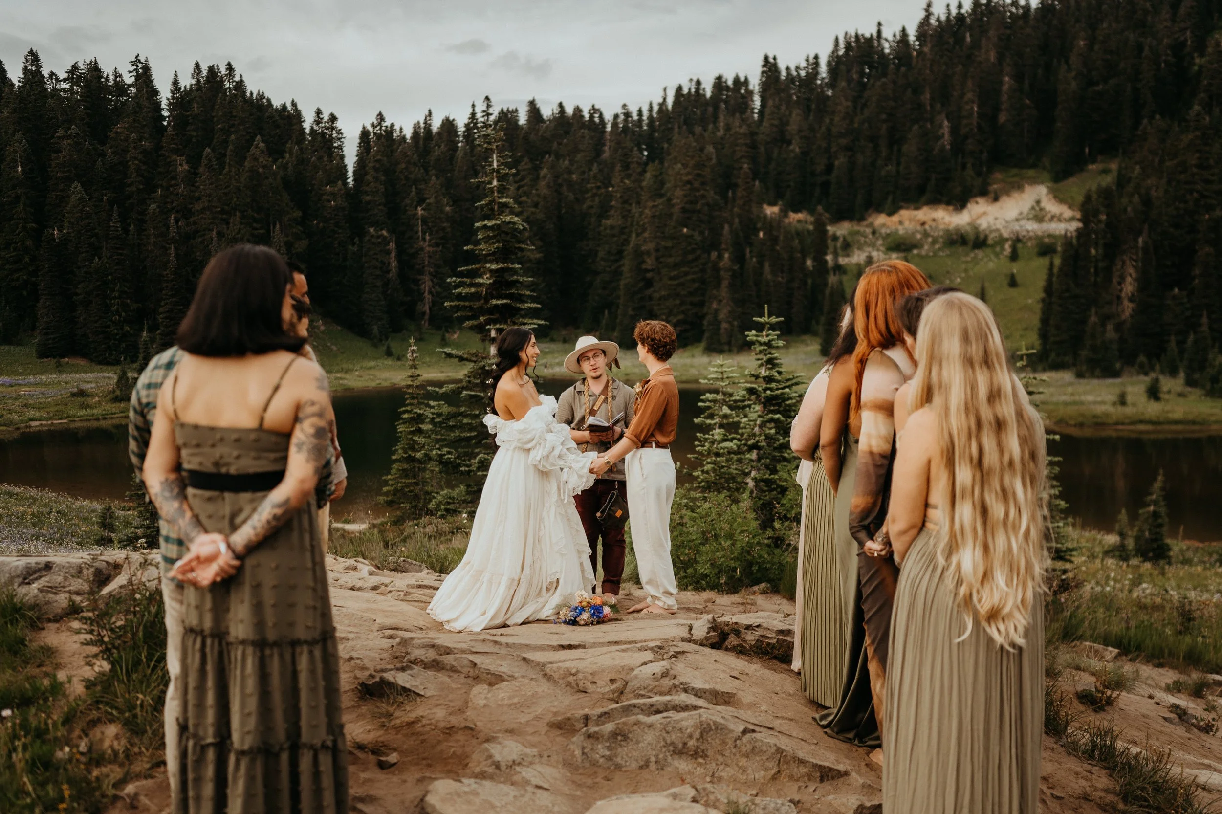 A group of people participating in a wedding ceremony outdoors by a lake, with a forested mountain background. The bride and groom are holding hands, while the officiant speaks. Several guests, some with long hairstyles, stand nearby.