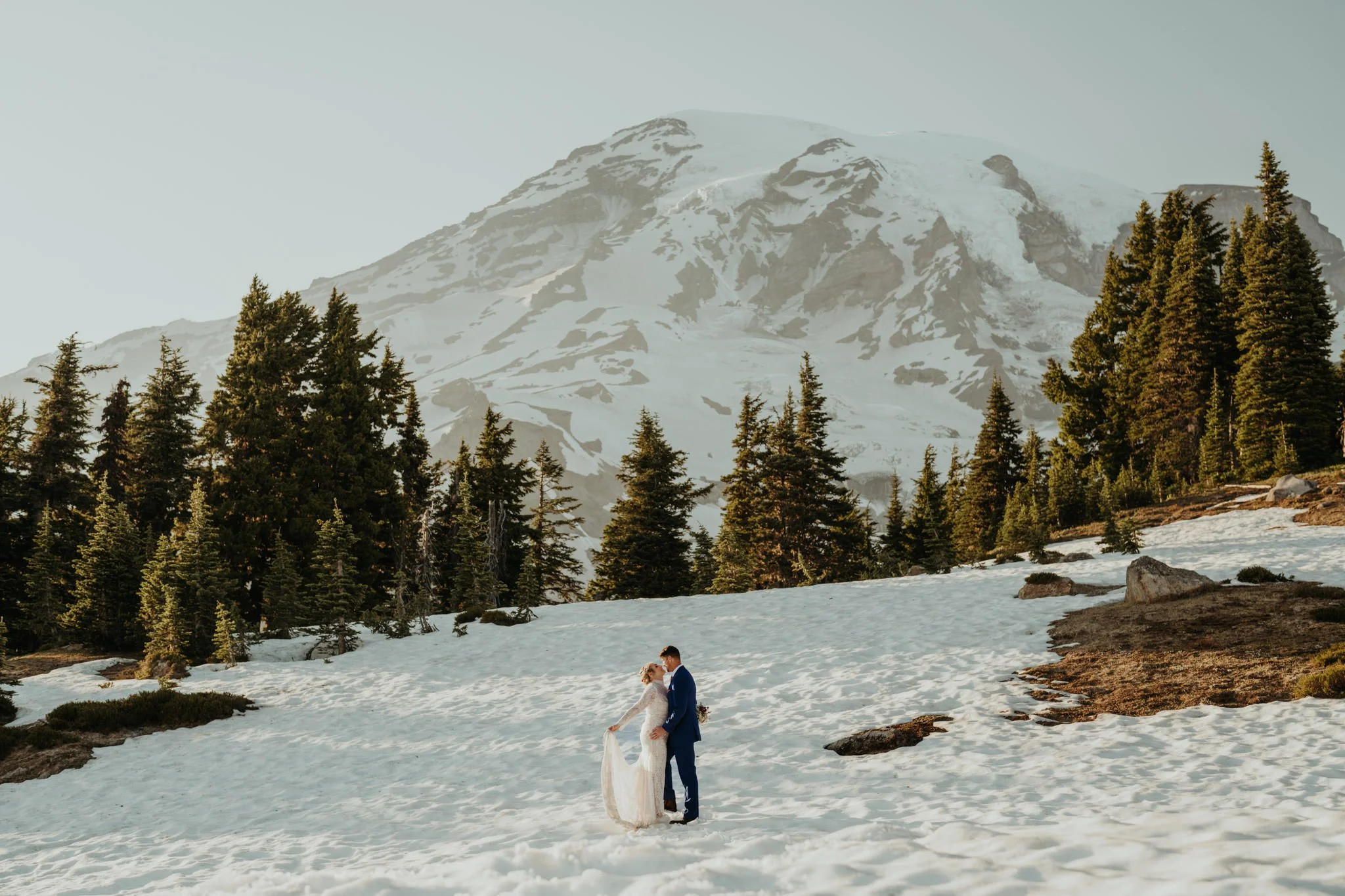 A bride and groom standing close together in a snowy landscape with a mountain and evergreen trees in the background.