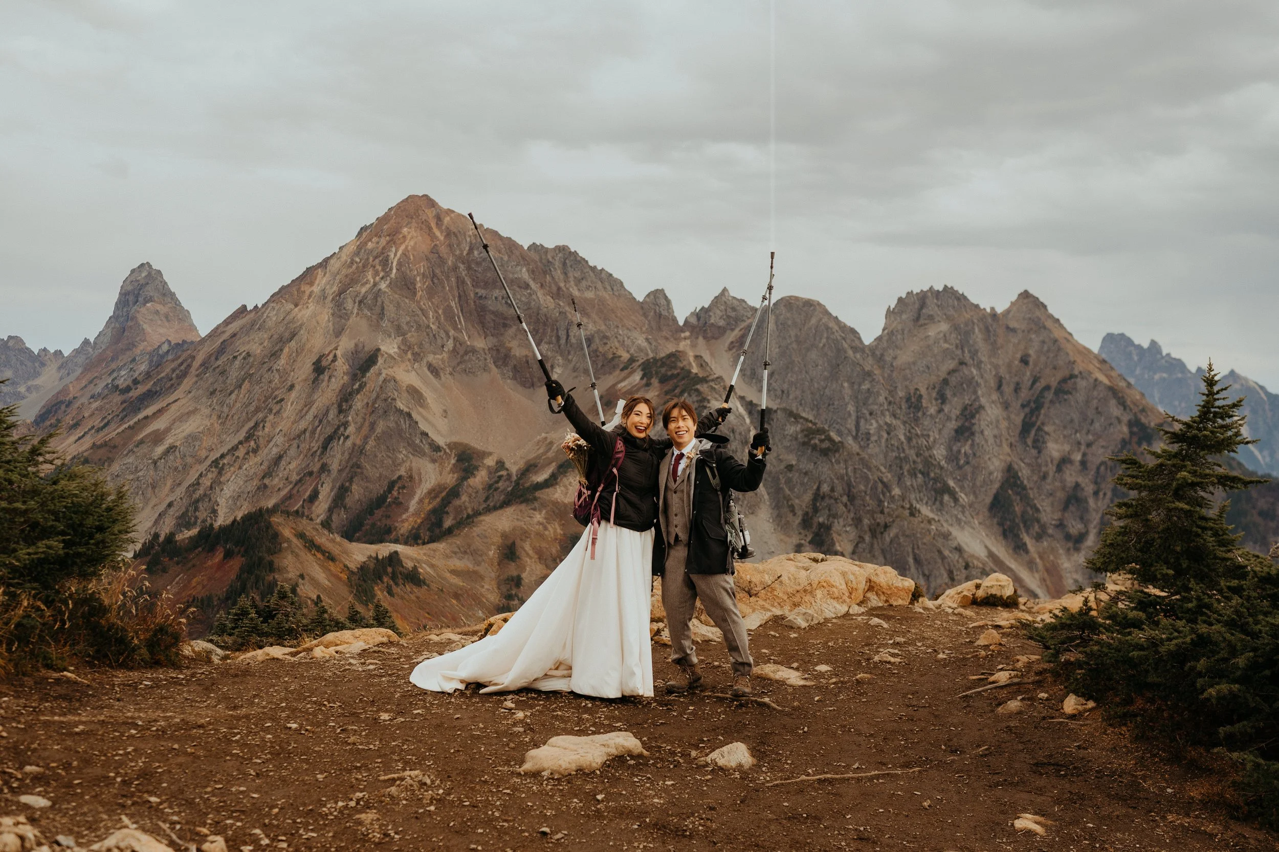 Two smiling people in wedding attire and hiking gear holding their trekking poles high, celebrating on a mountain trail with rugged peaks in the background.