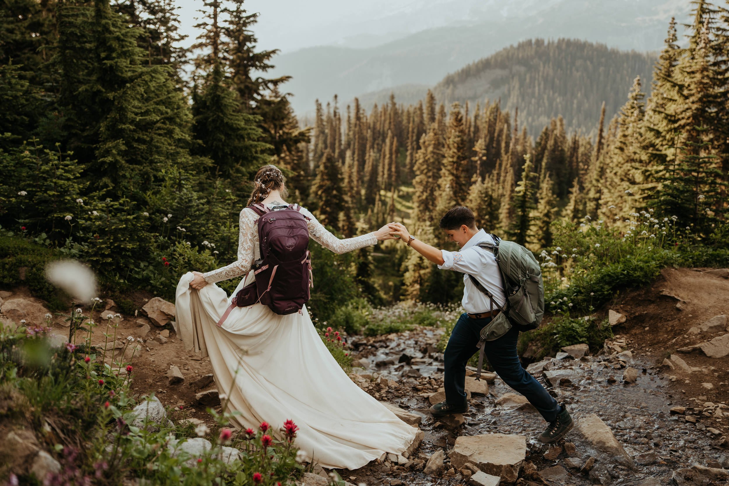 A bride in a white wedding dress holding hands with a bride in a white shirt and dark pants as she helps her cross a rocky stream in a forested mountain area.